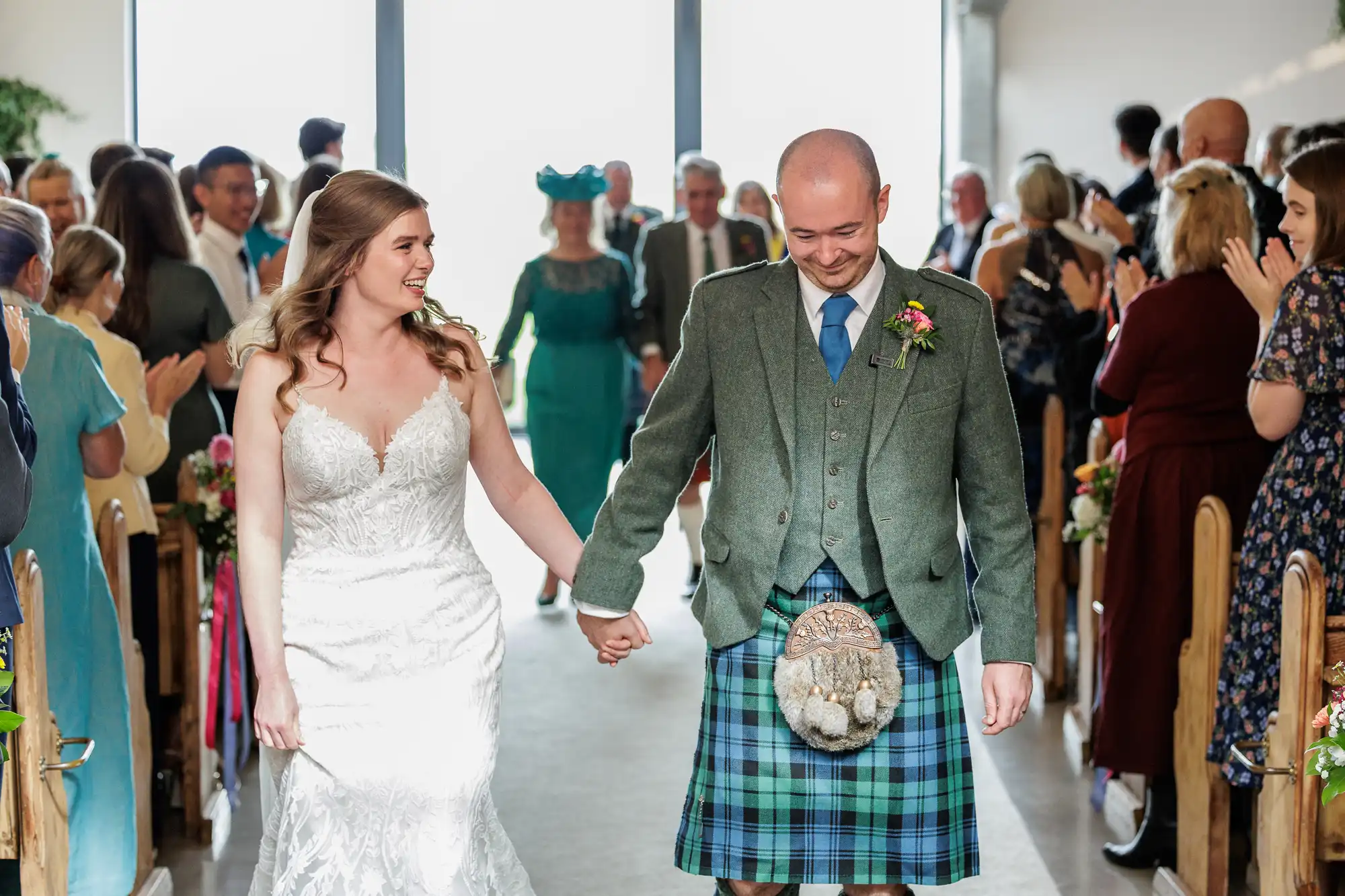 A bride in a white lace dress and a groom in a green jacket with a blue tartan kilt hold hands and smile whilst walking down the aisle. Guests on both sides, some applauding, watch the couple. Bright light streams in from tall windows behind them.