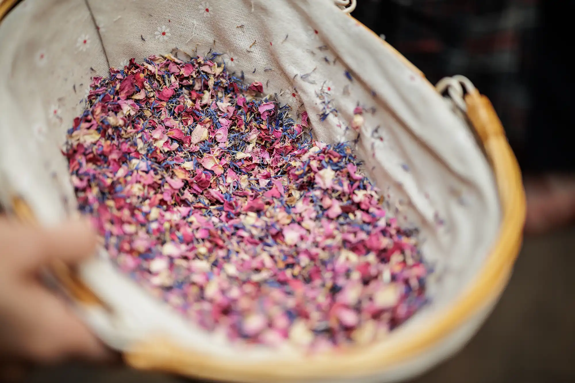 A close-up of a fabric-lined basket filled with colourful dried flower petals in shades of pink, purple, and blue. A hand holds the basket, and the background is blurred. The petals appear delicate, creating a vibrant, textured mixture.