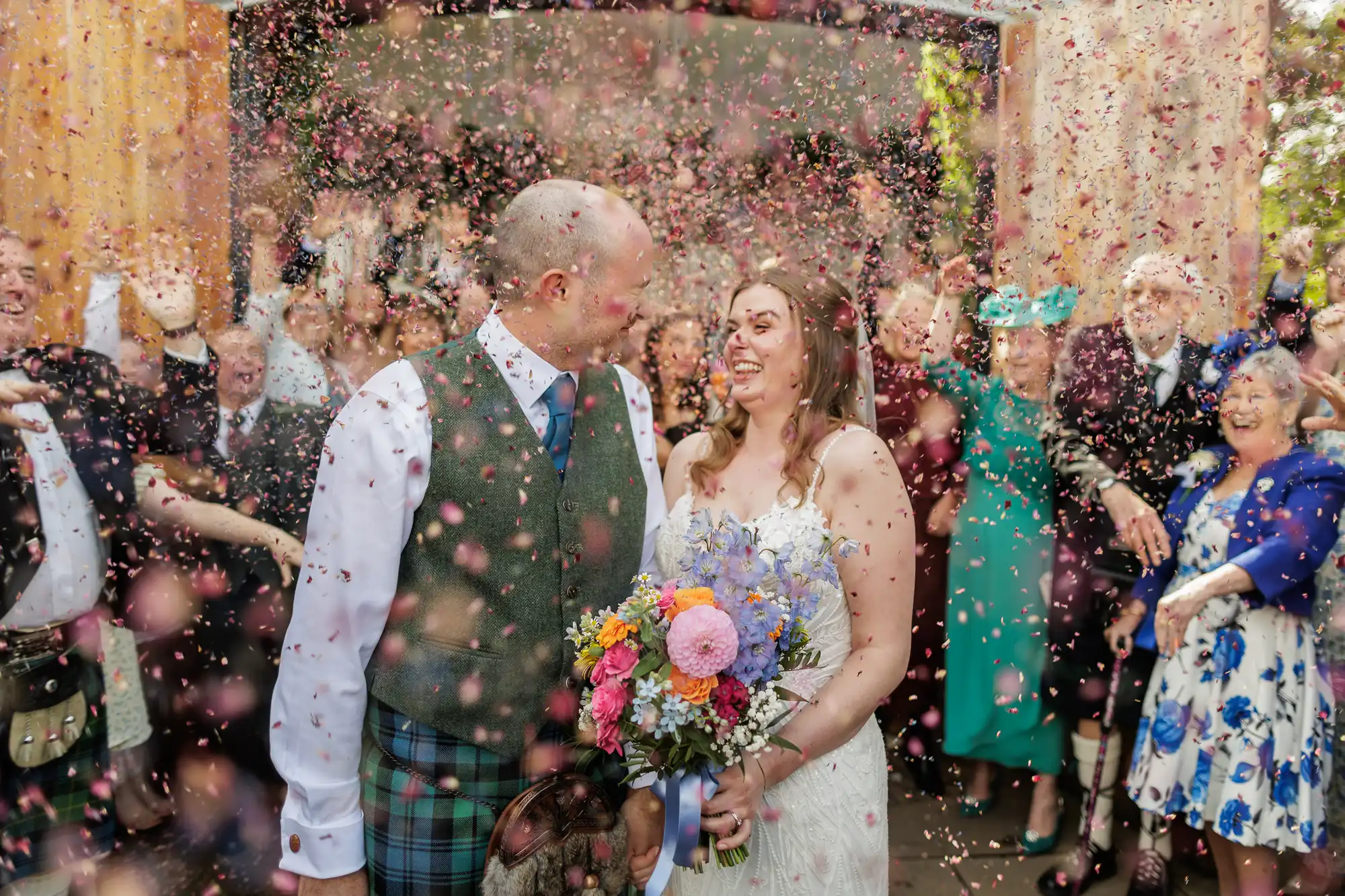 A newlywed couple smile at each other as guests shower them with flower petals. The groom wears a Scottish kilt outfit, and the bride holds a colourful bouquet. People in festive attire cheer in the background, celebrating the joyous moment.
