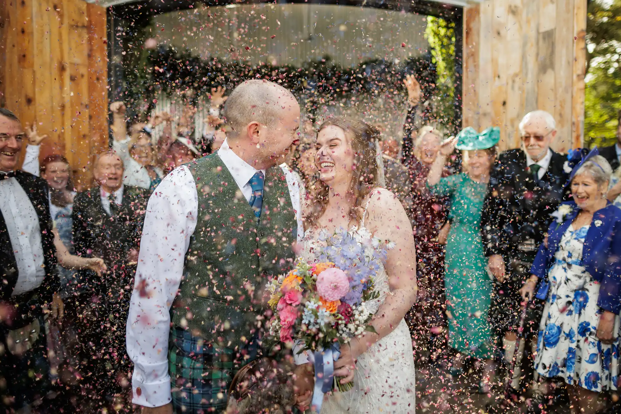 A newlywed couple, smiling and holding hands, stand amid a shower of colourful confetti. The bride holds a bouquet of flowers, and guests behind them cheer with raised arms. The joyful scene takes place outdoors, framed by a wooden archway.
