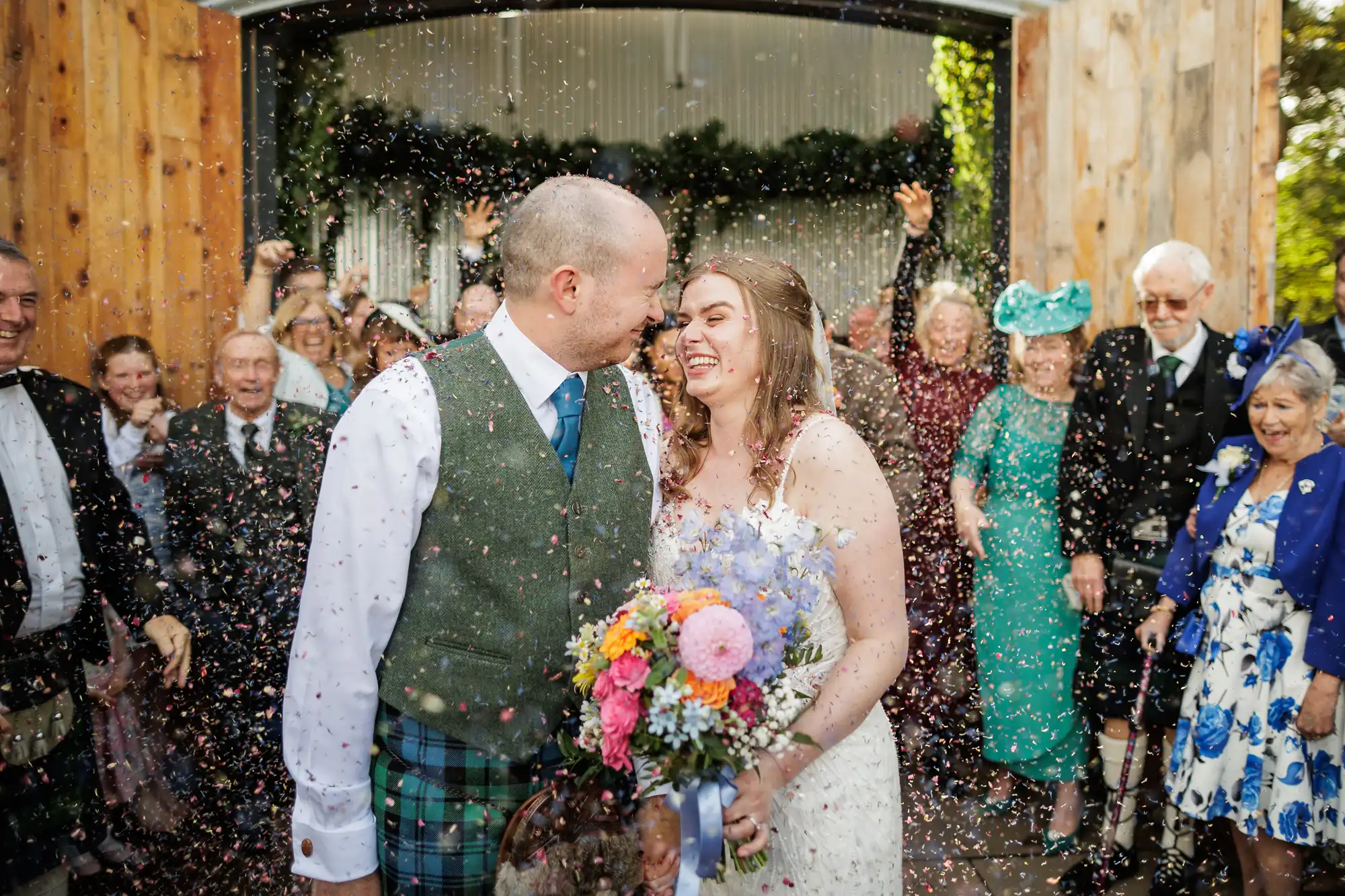 A bride and groom smile at each other whilst guests throw confetti. The groom wears a green waistcoat and tartan kilt; the bride wears a white dress and holds a colourful bouquet. Guests in festive attire cheer and celebrate in the background.