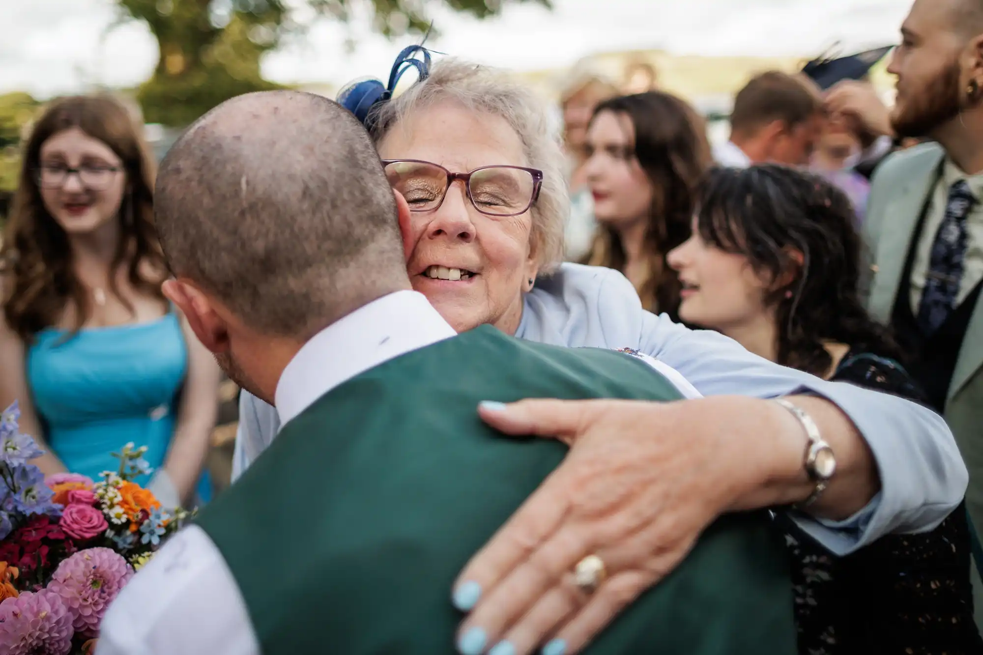 An elderly woman in spectacles warmly hugs a man wearing a green waistcoat, smiling with eyes closed. They are surrounded by people in formal attire, some holding colourful flowers, at an outdoor gathering under daylight.