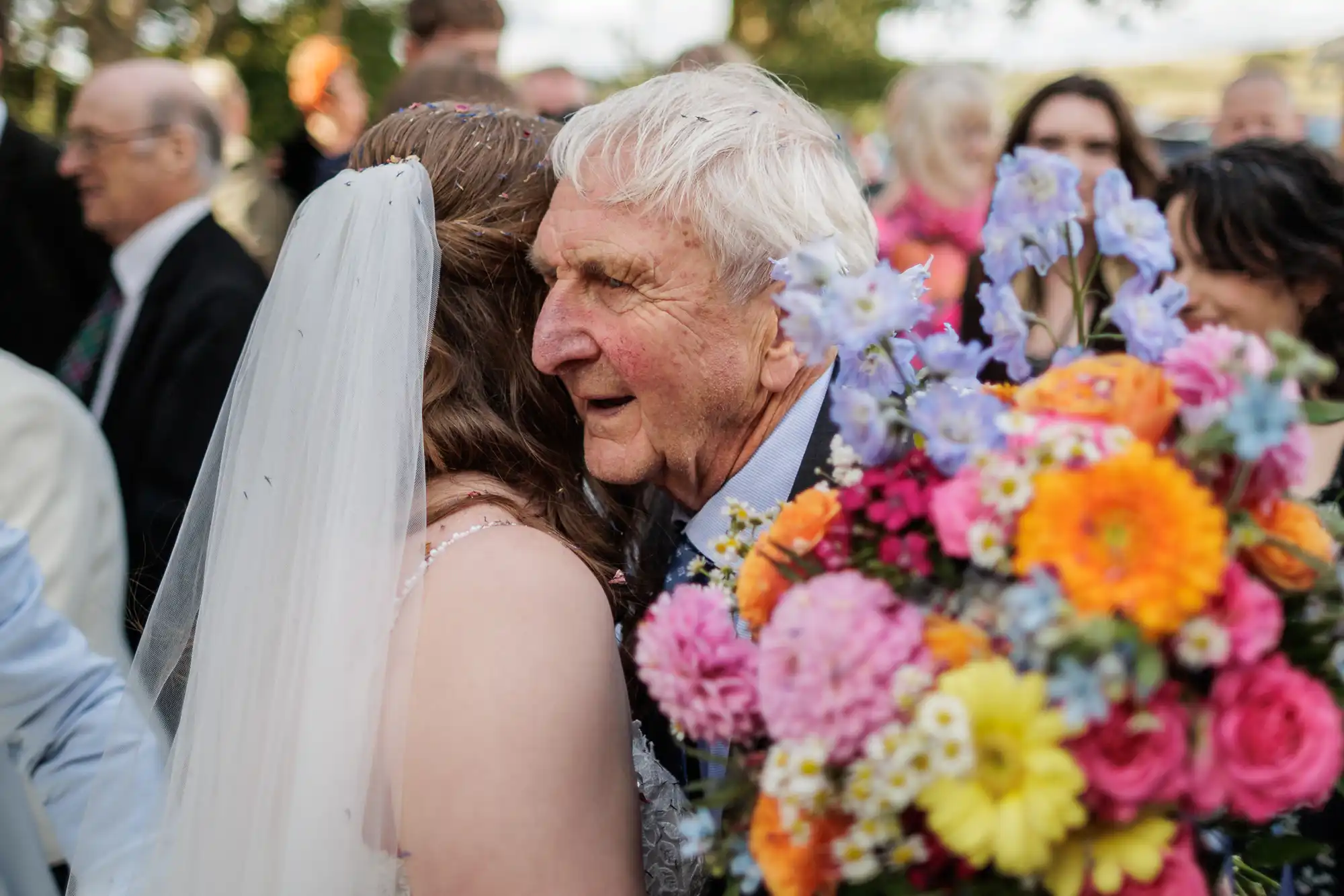 An elderly man with white hair hugs a bride in a white dress and veil. The bride holds a large, colourful bouquet of flowers. The scene is outdoors, with blurred people and greenery in the background, creating a joyful, celebratory atmosphere.