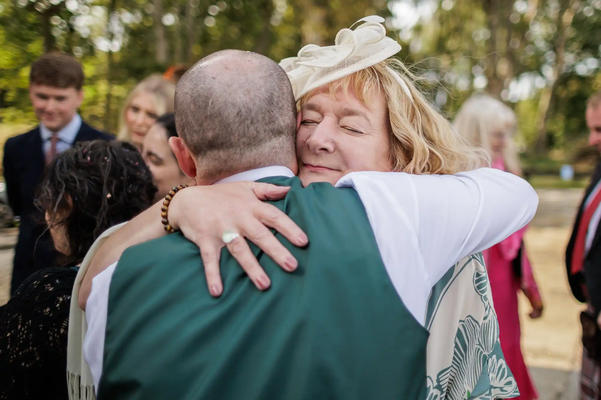 A woman with blonde hair in a decorative cream hat hugs a man in a green waistcoat outdoors. She smiles with her eyes closed. Several people in formal attire stand blurred in the background among trees. It appears to be a joyful occasion.