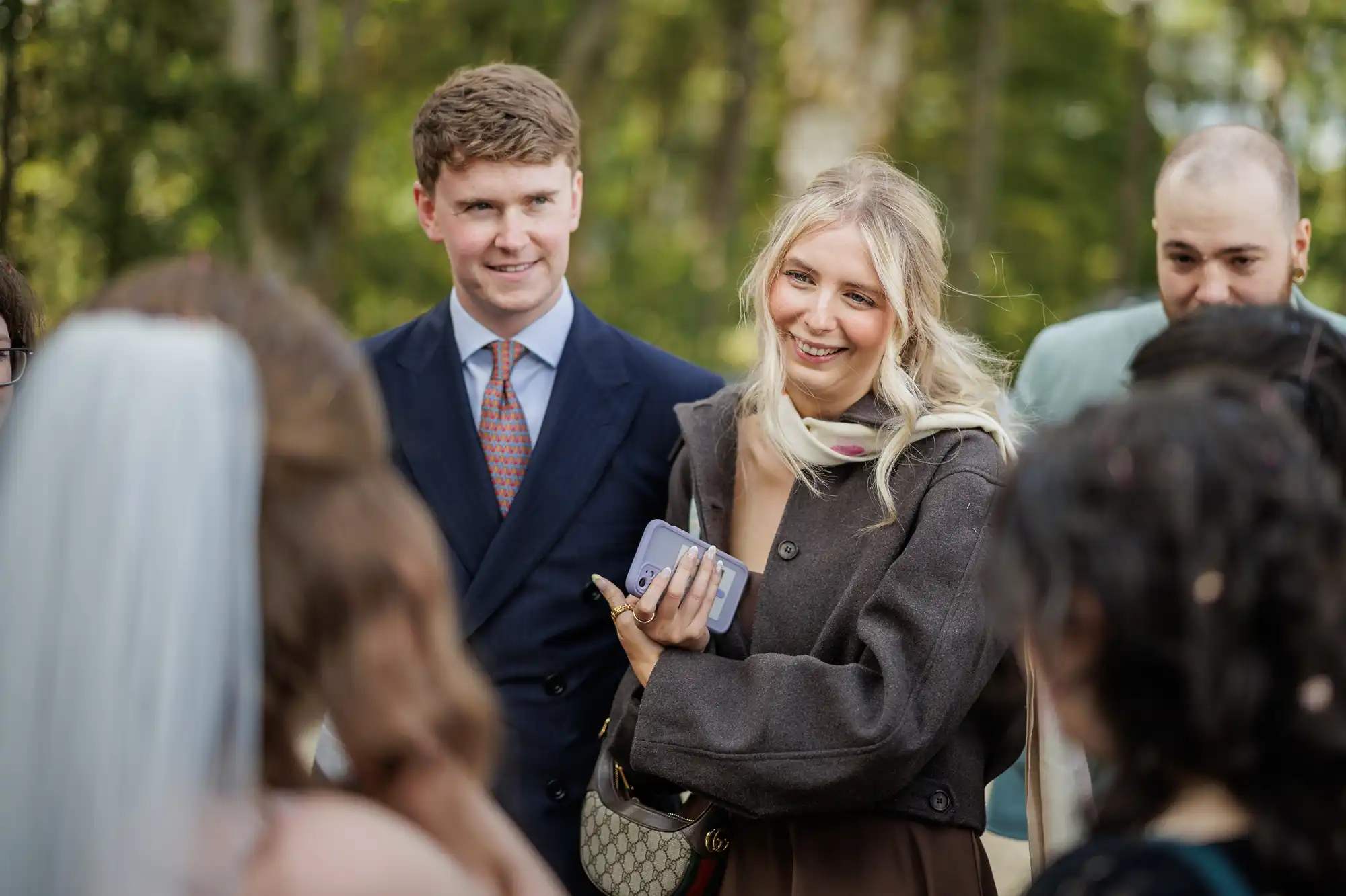 A young woman, smiling and holding a mobile, stands outdoors among a small group. Next to her, a man in a suit looks on. The group appears to be in a park or garden, with blurred greenery in the background. A bride in a veil is partly visible in the foreground.