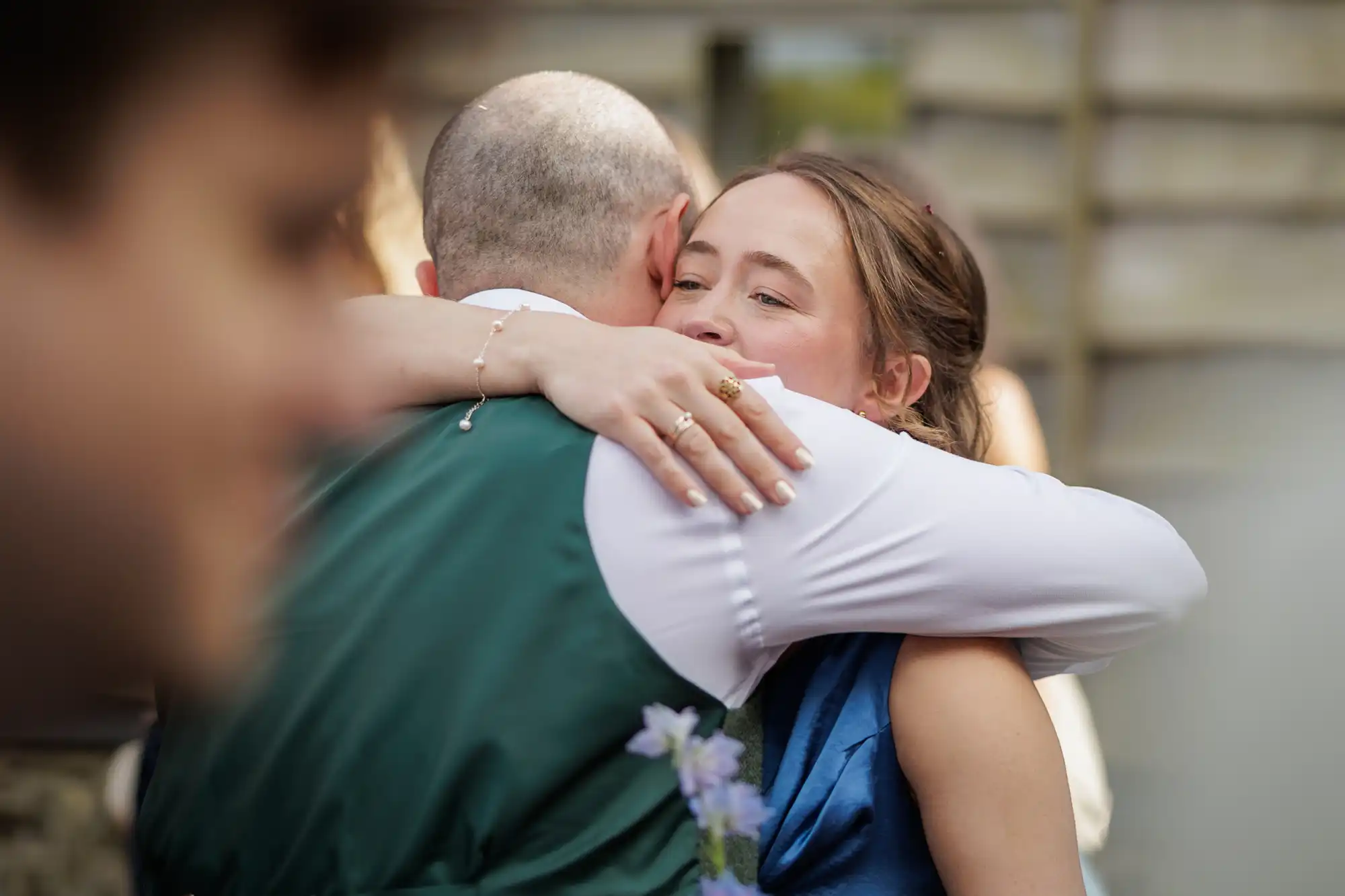 A woman in a blue dress warmly hugs a man in a green waistcoat and white shirt. Her face shows a joyful smile, and her hand rests on his back. The background is softly blurred, with another person slightly visible in the foreground.