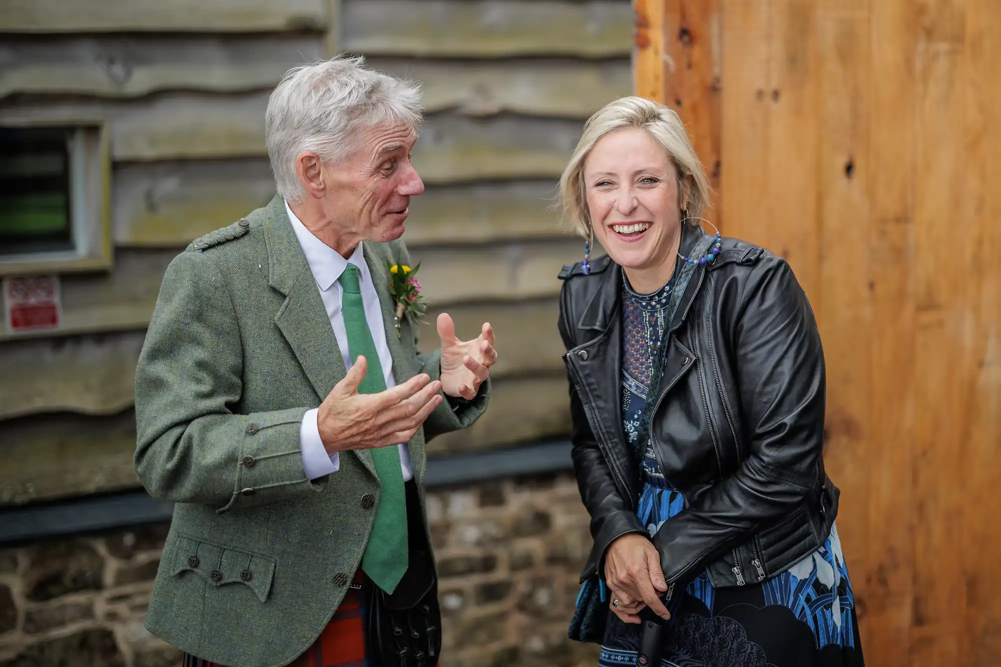 An older man in a green suit jacket and tie gestures as he talks to a smiling blonde woman in a patterned dress and dark jacket. They stand outdoors near a wooden building, both appearing happy and engaged in conversation.