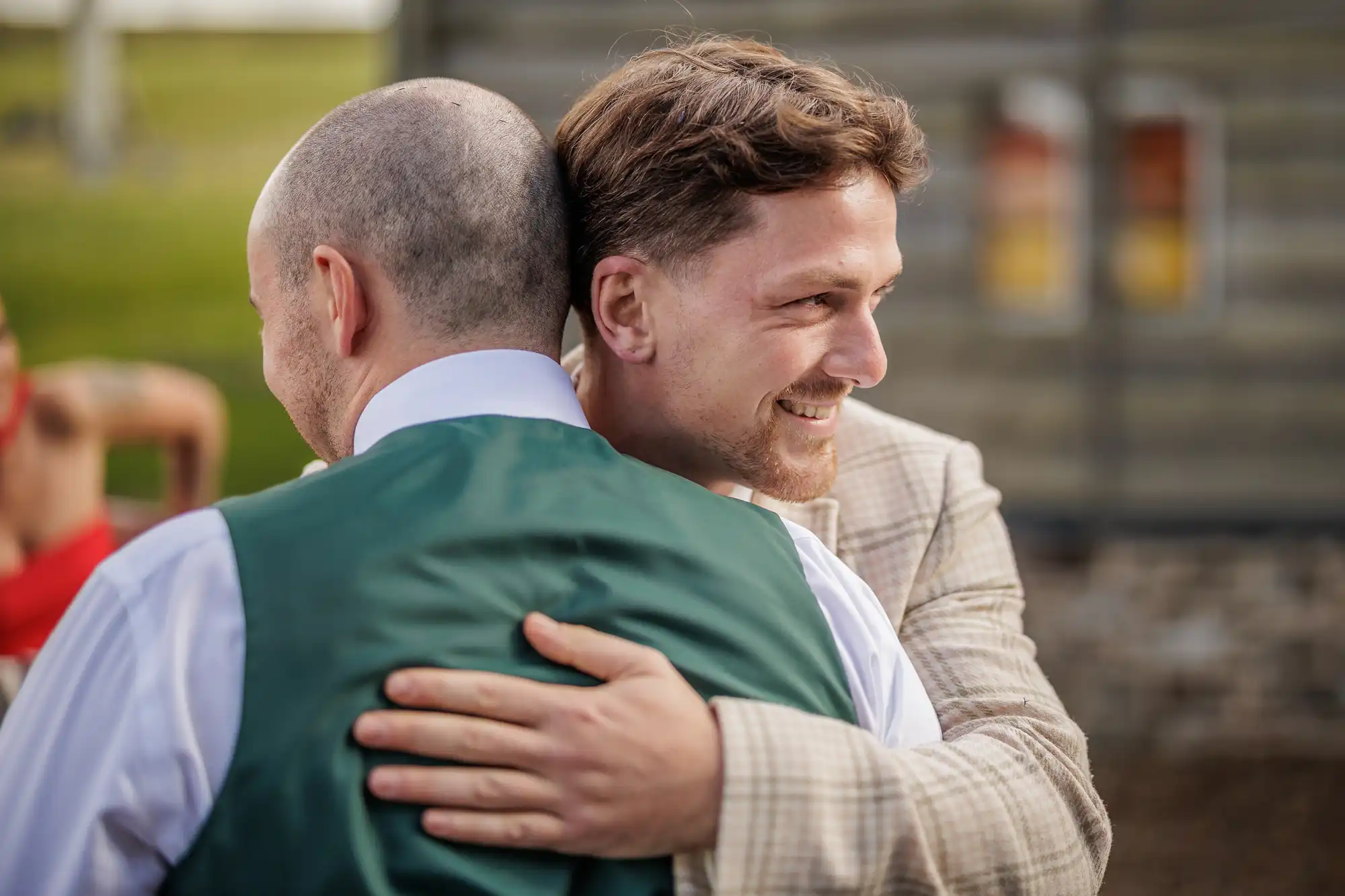 Two men hug outdoors. One faces away, wearing a white shirt and green waistcoat. The other, in a light checked jacket, smiles warmly with his arm around the first man's back. A blurred building and greenery appear in the background.