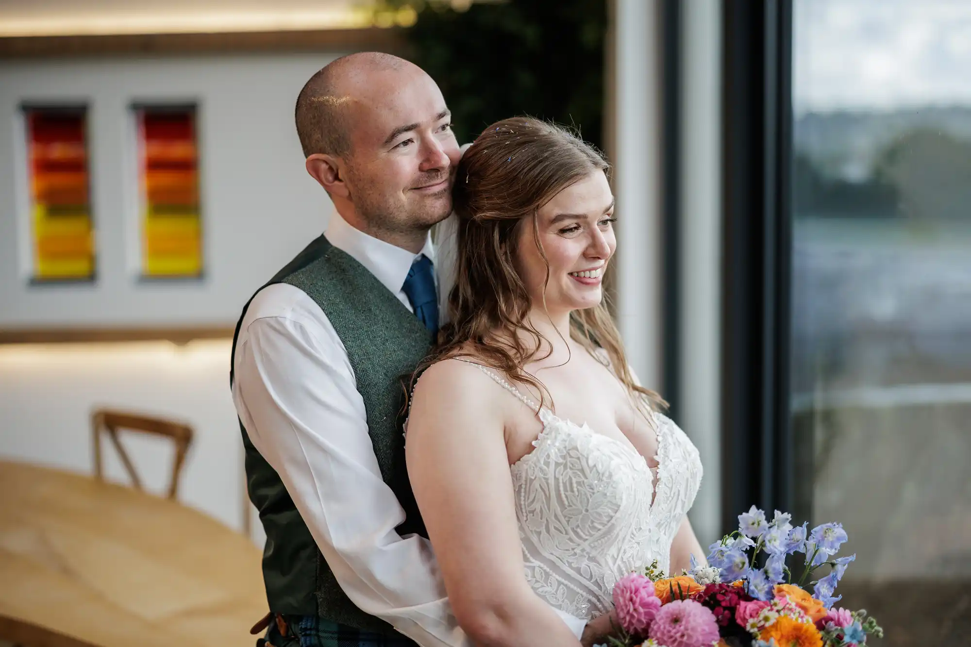 A smiling bride in a white lace dress holds a colourful bouquet while her groom, in a green waistcoat and tie, lovingly hugs her from behind. They stand indoors by a large window, with modern art and a wooden table in the background.