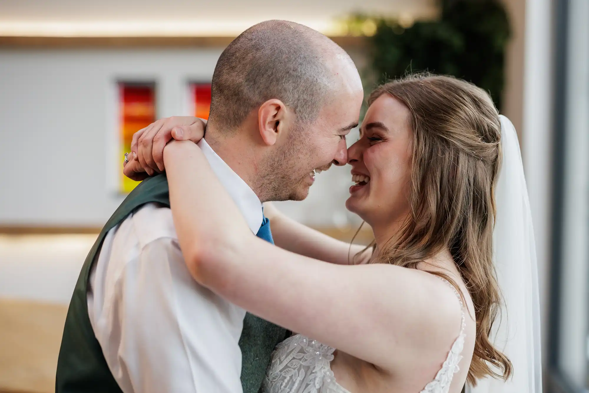 A smiling bride and groom embrace, foreheads touching and eyes closed, sharing a joyful moment. The bride wears a white dress and veil, while the groom wears a white shirt, waistcoat, and blue tie. The background is softly lit with colourful abstract art on the wall.