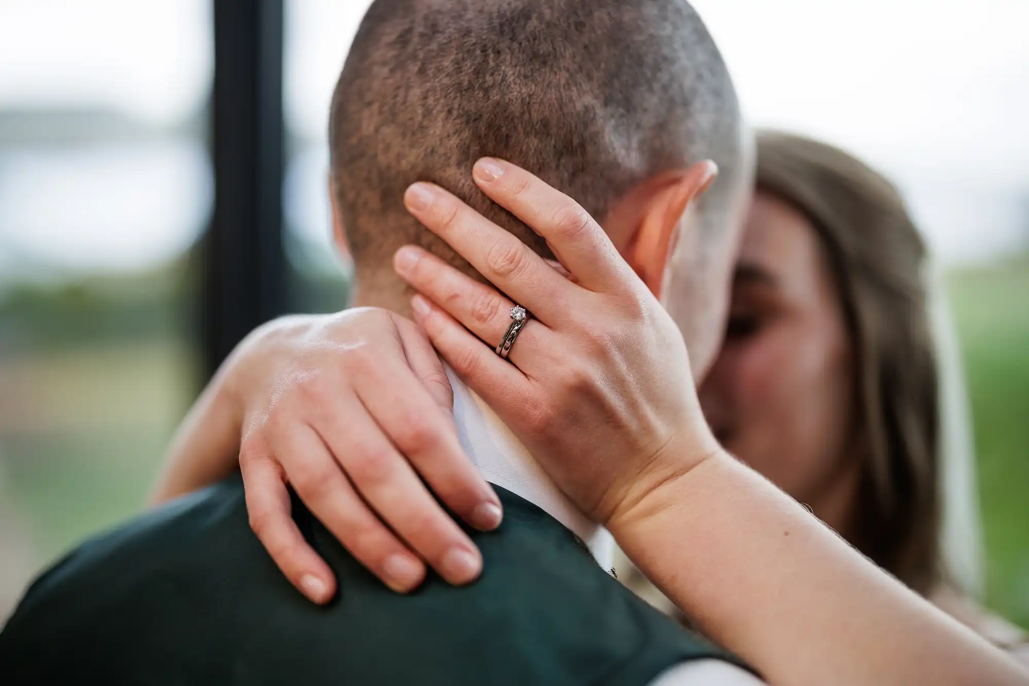 A close-up of a couple embracing, with the woman’s hands around the man’s neck. She wears a diamond engagement ring and wedding band. The focus is on her hands and rings, while their faces are blurred in an intimate, tender moment.