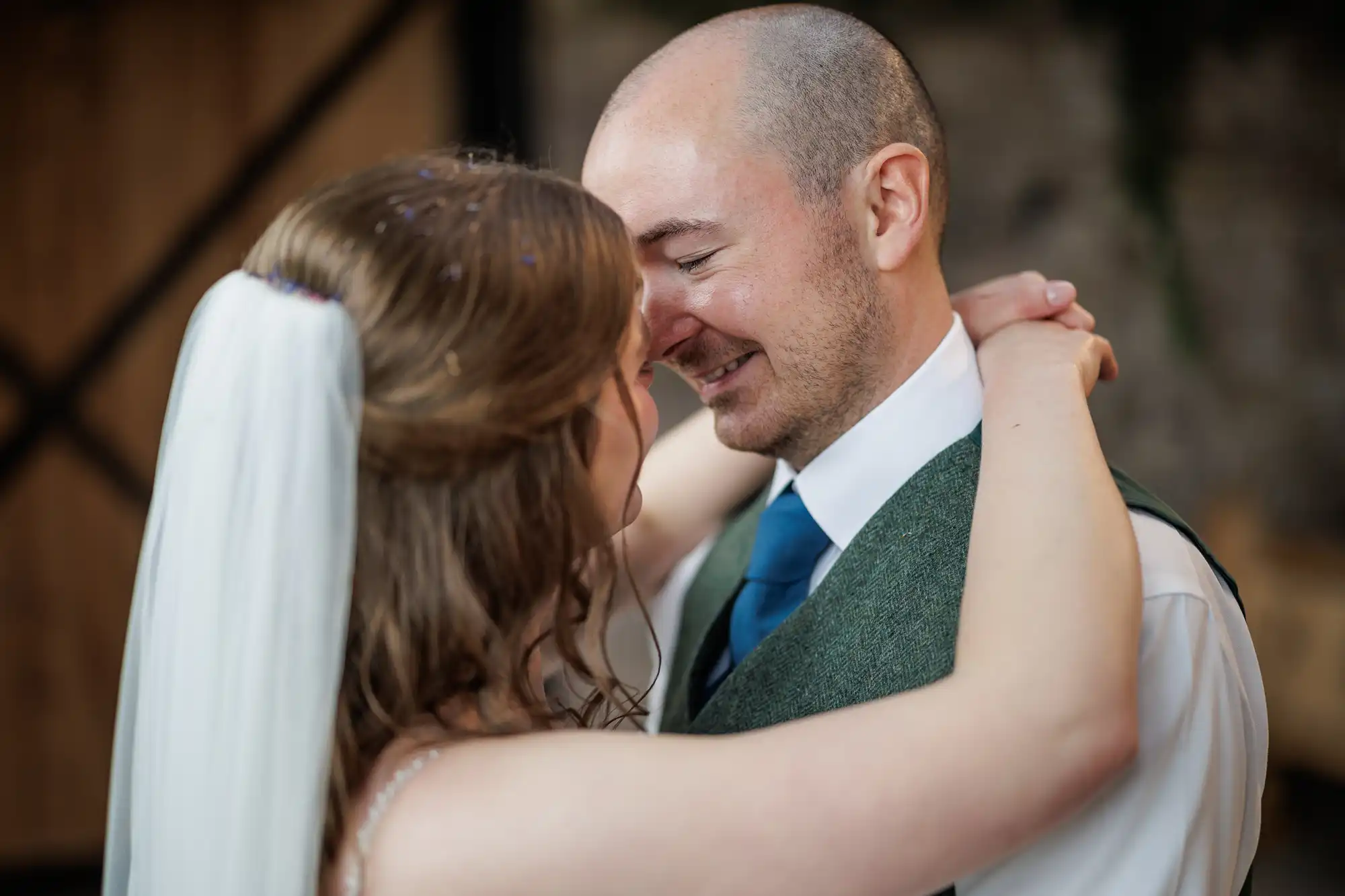 A bride in a white dress and veil embraces a smiling groom in a green waistcoat, white shirt, and blue tie. They look into each other's eyes, sharing an intimate moment, with their arms around each other in a warmly lit indoor setting.
