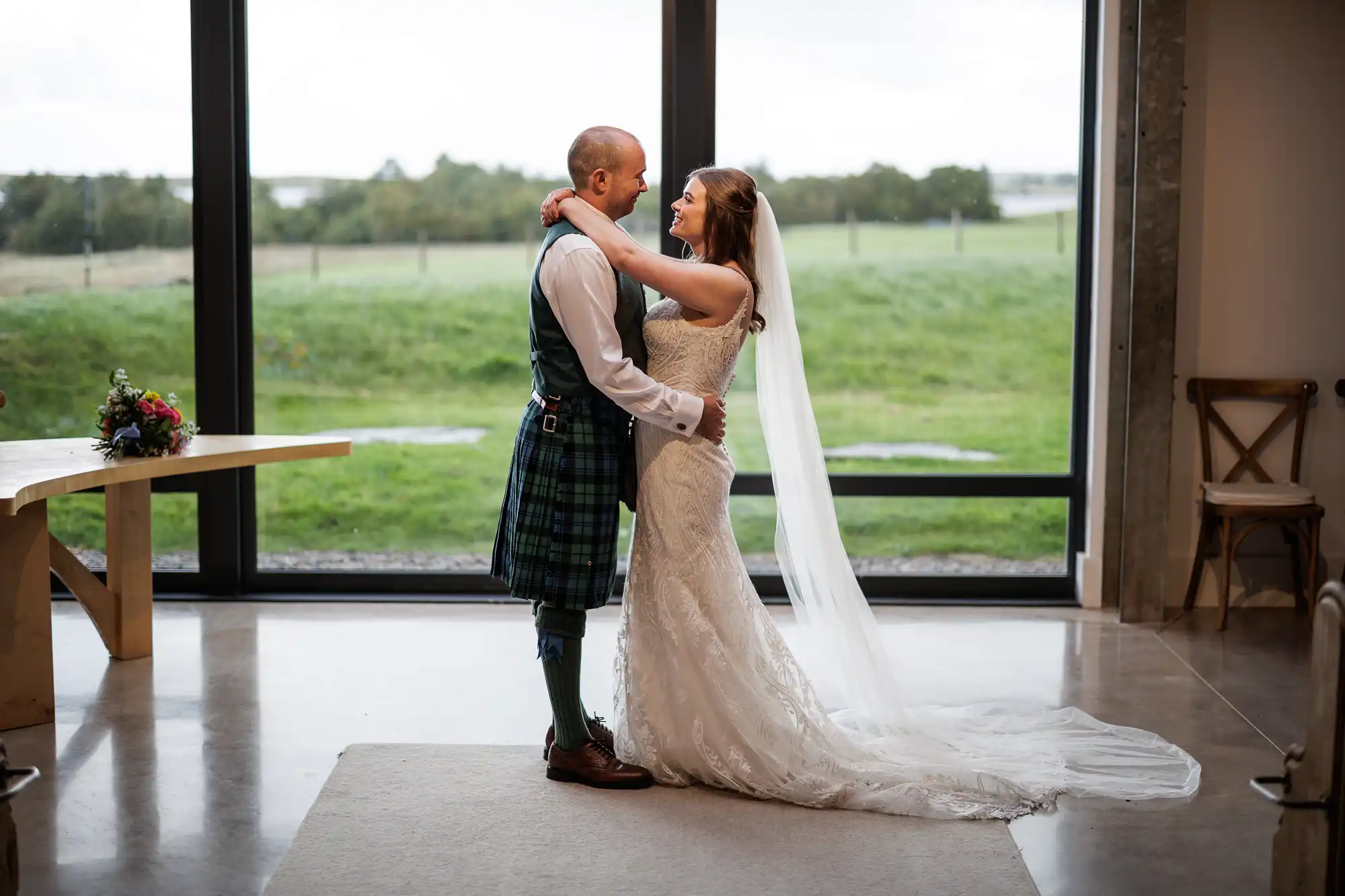 A bride in a white lace gown and long veil embraces a groom wearing a traditional Scottish kilt. They face each other, smiling, indoors by large windows with a scenic green field and cloudy sky visible outside. A bouquet sits on a nearby table.