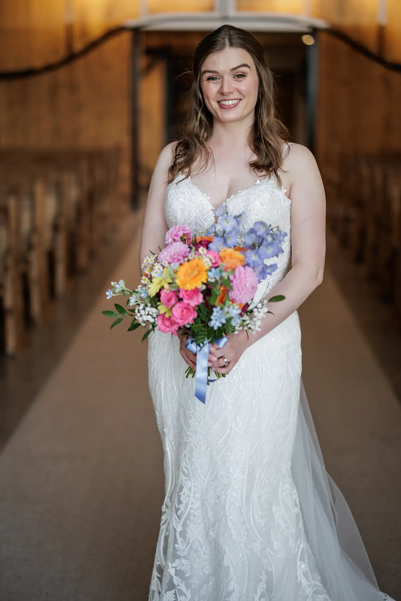 A smiling bride stands indoors, wearing a white lace wedding dress with thin straps. She holds a colourful bouquet of flowers including pink, purple, yellow, and orange blooms. The background shows wooden pews and warm lighting.
