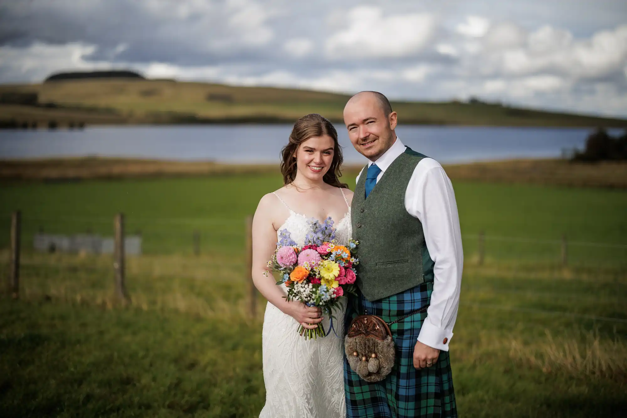 A smiling bride in a white dress holding a colourful bouquet stands beside a groom in a green waistcoat, white shirt, and blue tartan kilt. They pose outdoors in front of green fields, a lake, and a cloudy sky.