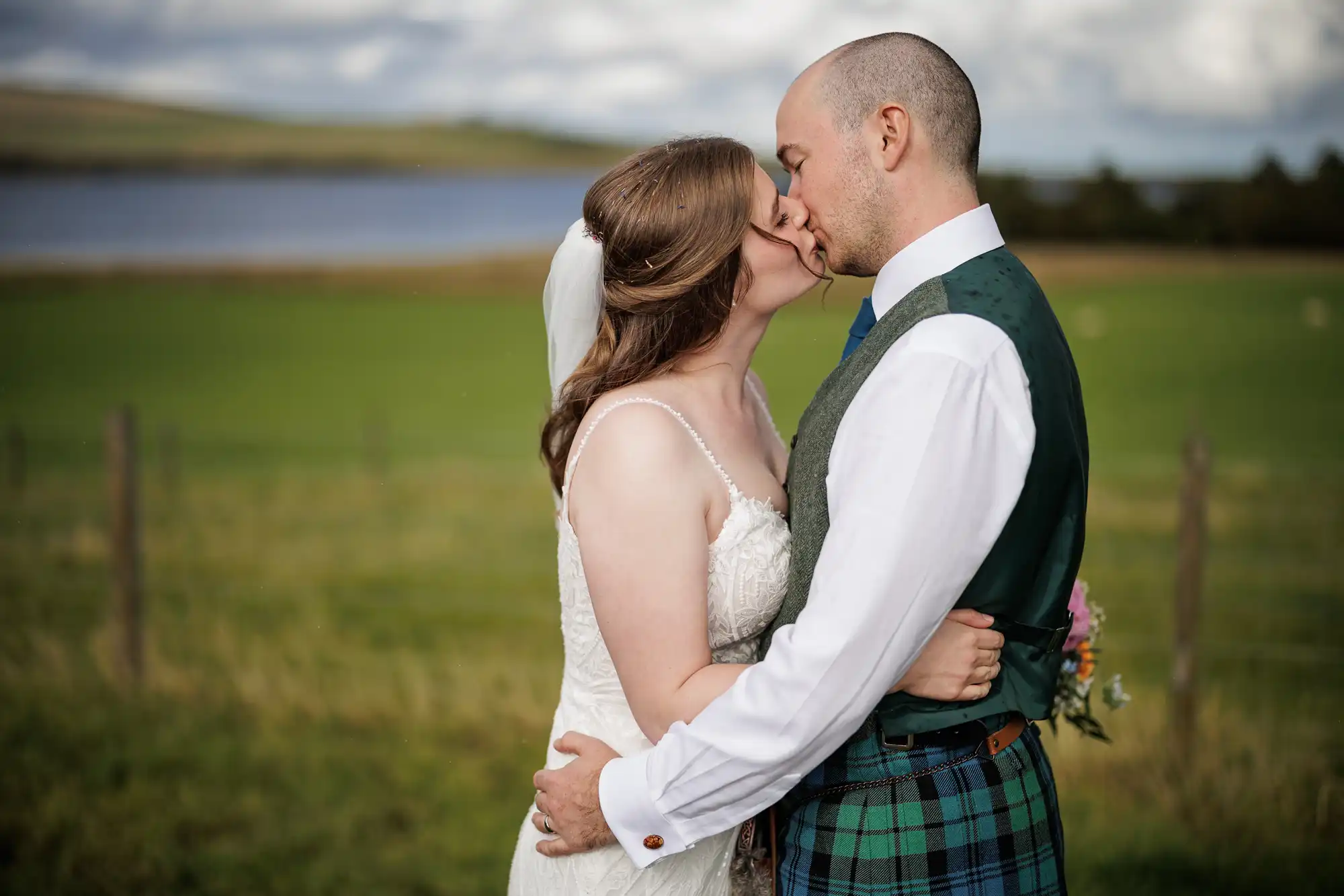 A bride in a white gown and veil kisses a groom dressed in a green tartan kilt and waistcoat. They embrace outdoors in front of a grassy field with a lake and hills in the blurred background, under a cloudy sky.