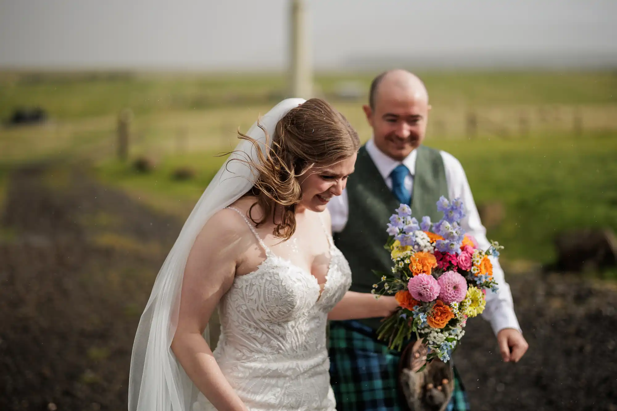 A bride in a white lace dress and veil smiles whilst holding a bouquet of colourful flowers. Next to her, a man in a blue checked kilt, green waistcoat, and tie also smiles. They are outdoors on a grassy path with a blurred background.