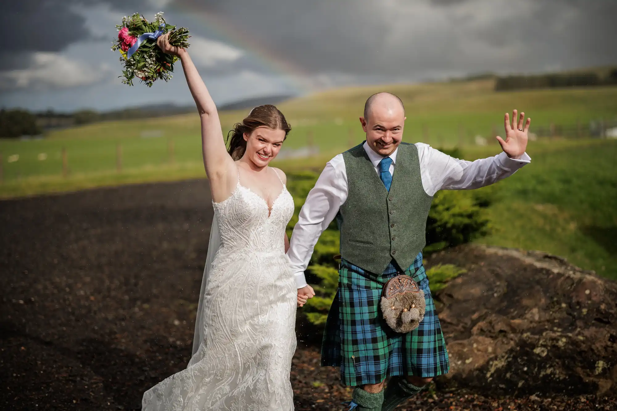 A joyful bride in a white lace dress holds a bouquet high, smiling and holding hands with a groom wearing a green waistcoat, blue tartan kilt, and sporran. They wave and walk outdoors on a cloudy day with green fields and a faint rainbow in the background.