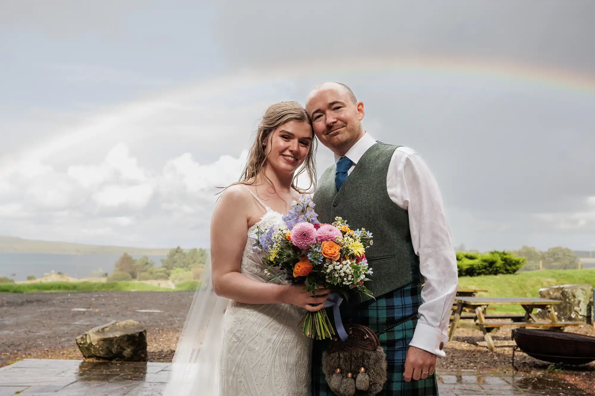 A bride in a white dress and veil stands beside a groom in a green waistcoat and blue tartan kilt. They smile, holding a colourful bouquet. A rainbow arcs across a cloudy sky in the background, with greenery and water visible behind them.