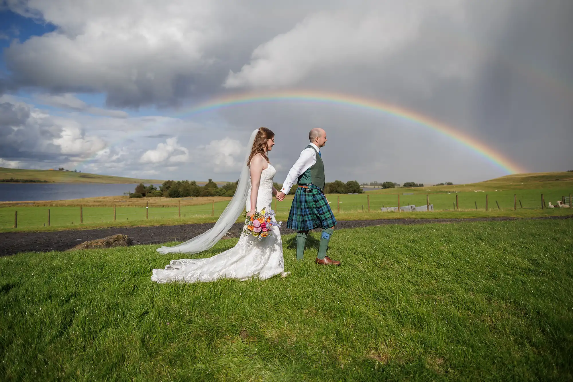 A bride in a white dress and veil holds hands with a groom wearing a kilt as they walk across green grass. A colourful bouquet is in the bride’s hand. A vivid rainbow arcs across a cloudy sky above the rural, open landscape in the background.