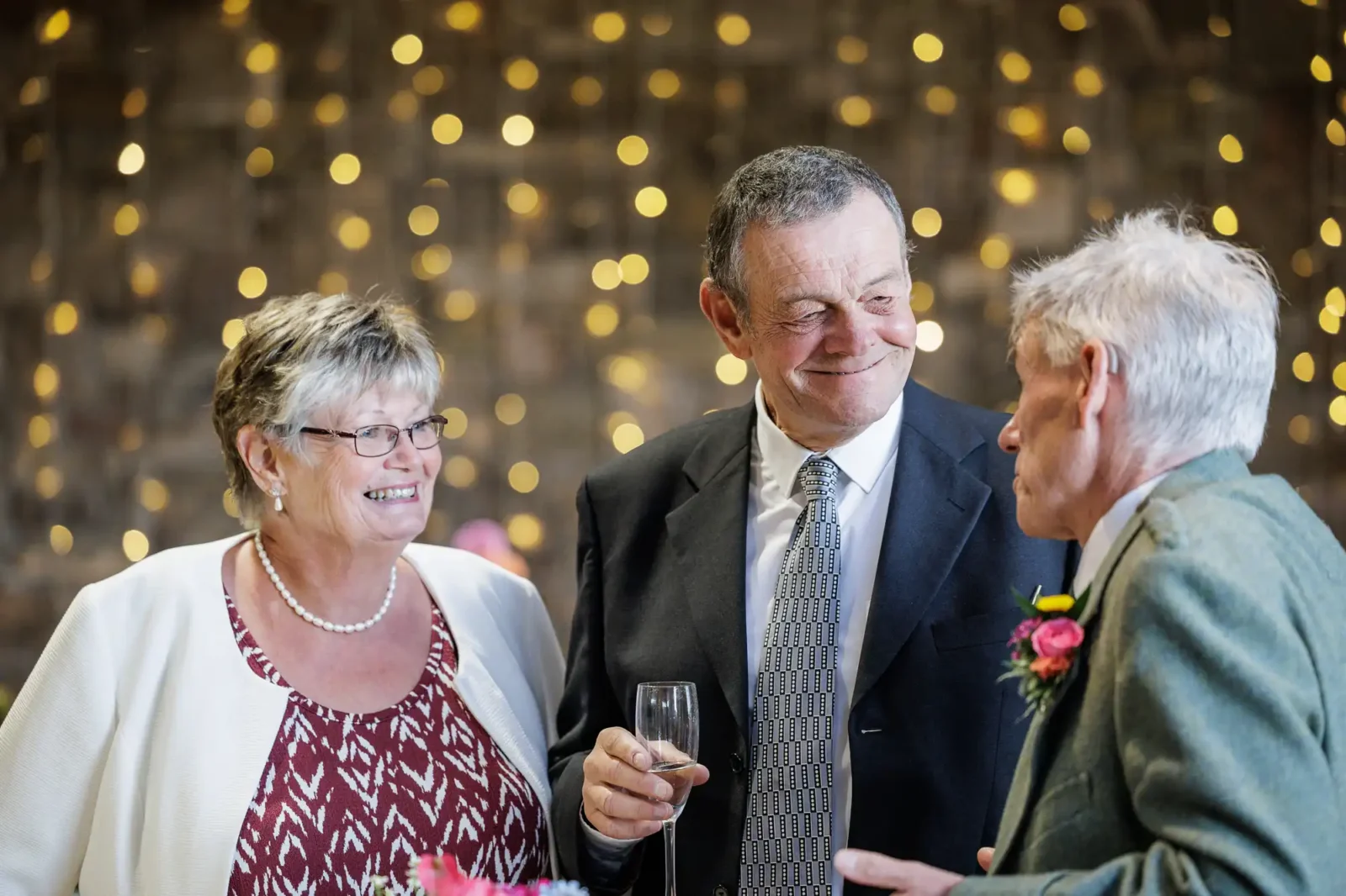 Documentary wedding photographer captures three older adults, two men in suits and a woman in glasses and a patterned dress with a white jacket, smile and converse at an event. One man holds a drink. Warm fairy lights blur in the background, creating a festive atmosphere.