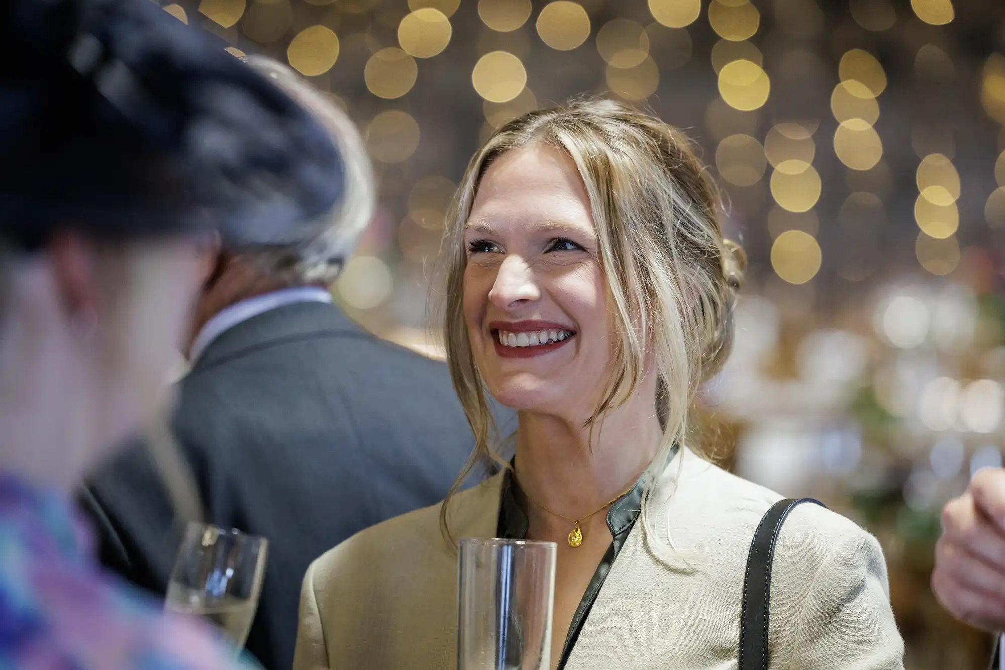 A woman with fair hair styled up smiles whilst holding a champagne flute at a social event. She wears a light-coloured jacket and a gold necklace. Out-of-focus people and warm, blurry fairy lights create a festive, elegant background.