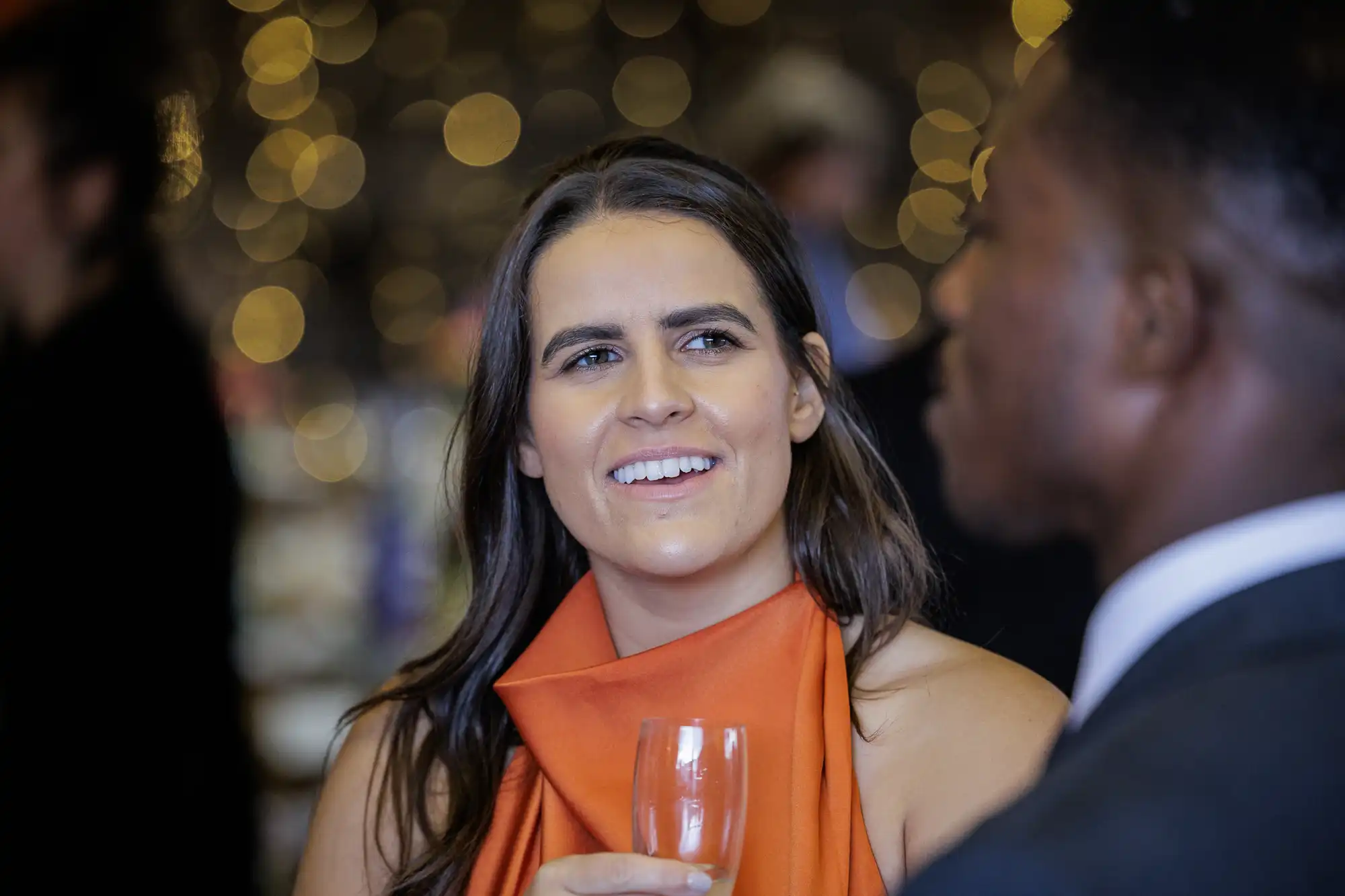 A woman in an orange dress smiles while holding a glass at an event. Soft golden lights blur in the background, creating a festive atmosphere. Another person in a dark suit is partially visible in the foreground, out of focus.