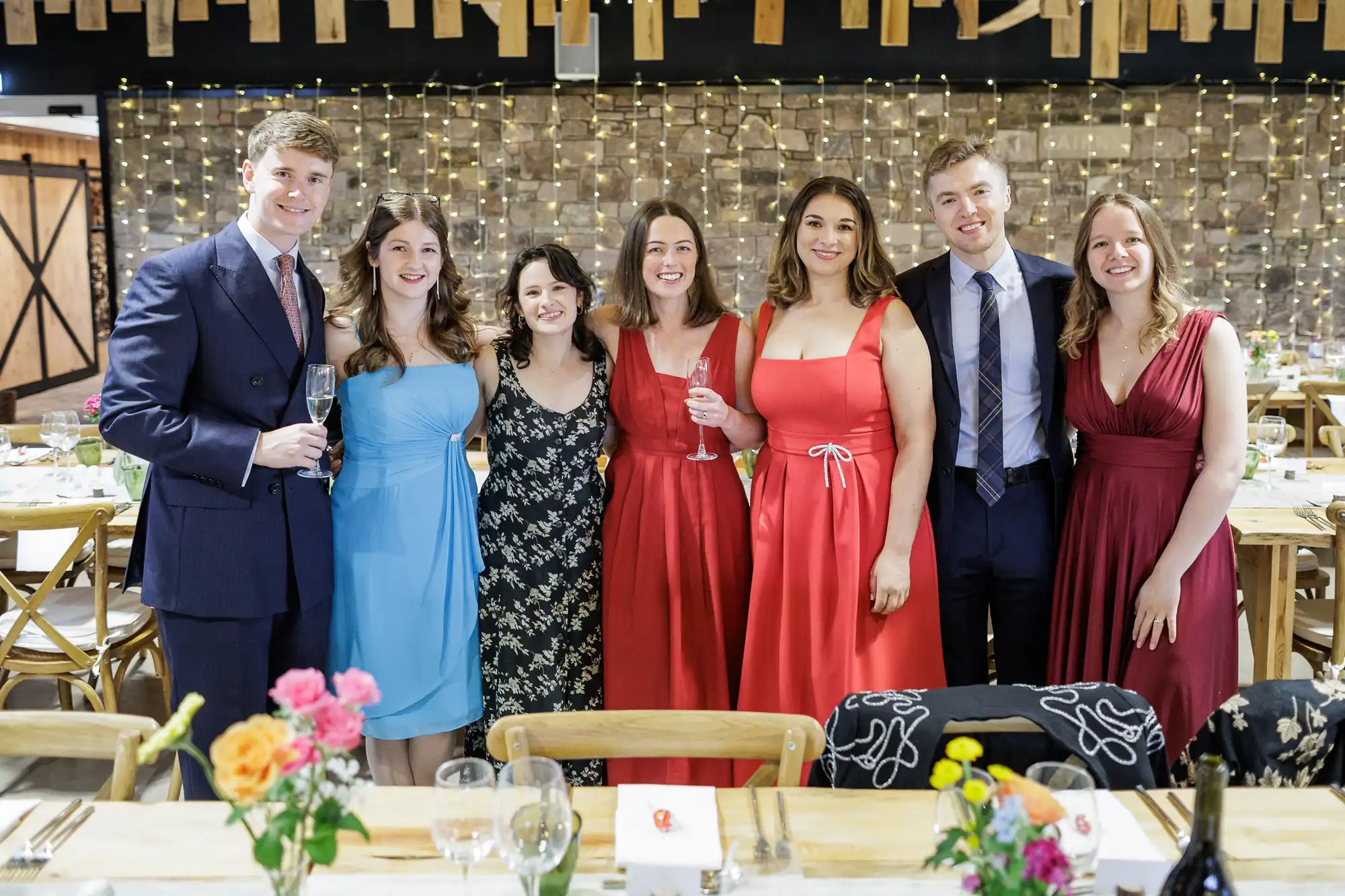 Seven young adults dressed formally pose together, smiling inside a warmly lit banquet hall decorated with fairy lights and floral centrepieces. Three women wear red or pink dresses, one wears blue, one wears a black floral dress, and the two men wear suits.