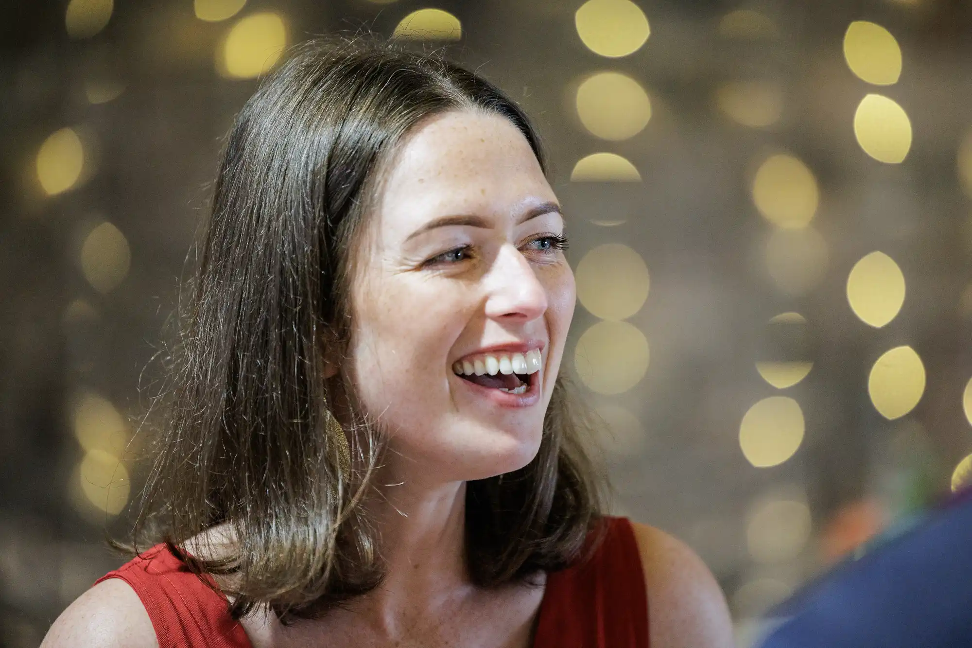 A woman with shoulder-length brown hair and wearing a sleeveless red top is smiling and laughing. The background is softly blurred with warm circular lights, creating a cheerful, festive atmosphere.