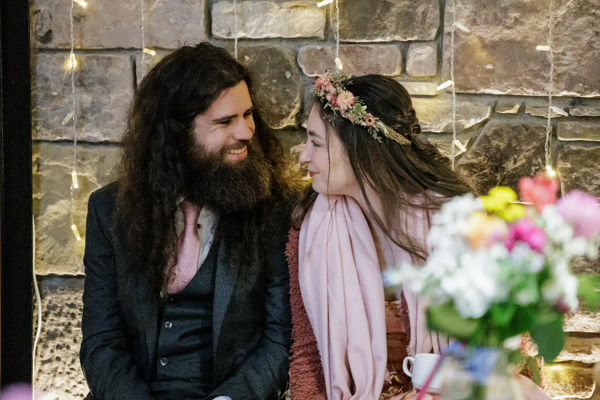 A man with long dark hair and a beard, wearing a suit, smiles at a woman with long hair and a flower crown, both sitting in front of a stone wall with fairy lights. A blurred bouquet of colourful flowers is in the foreground.