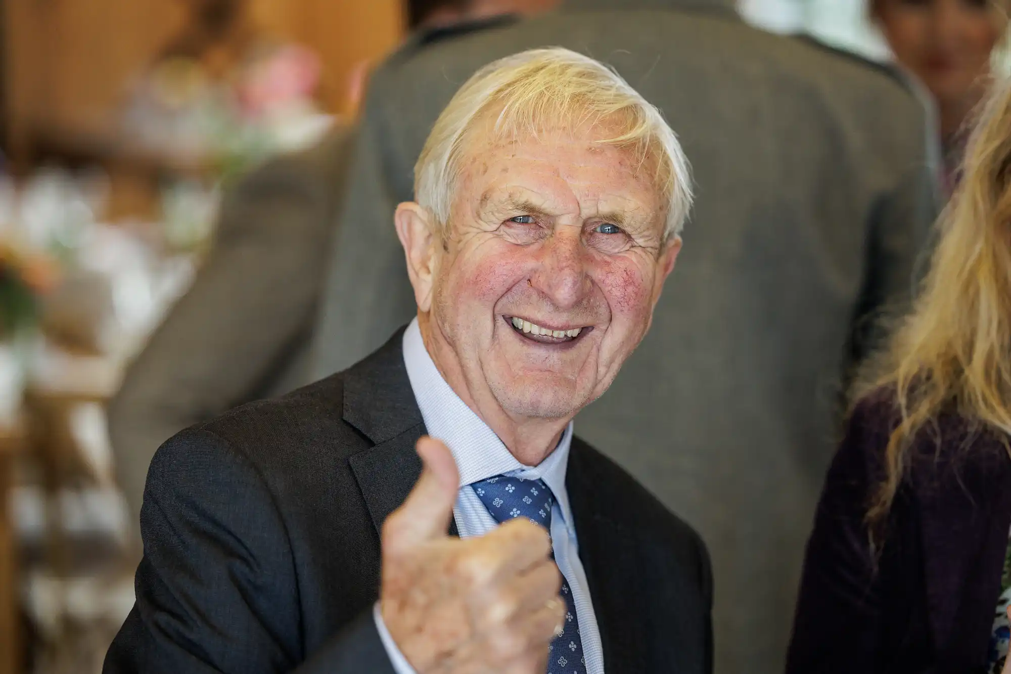 An elderly man with short white hair, wearing a dark suit and blue patterned tie, smiles warmly and gives a thumbs up. The background is softly blurred, showing people and a festive indoor setting, suggesting a celebratory event.