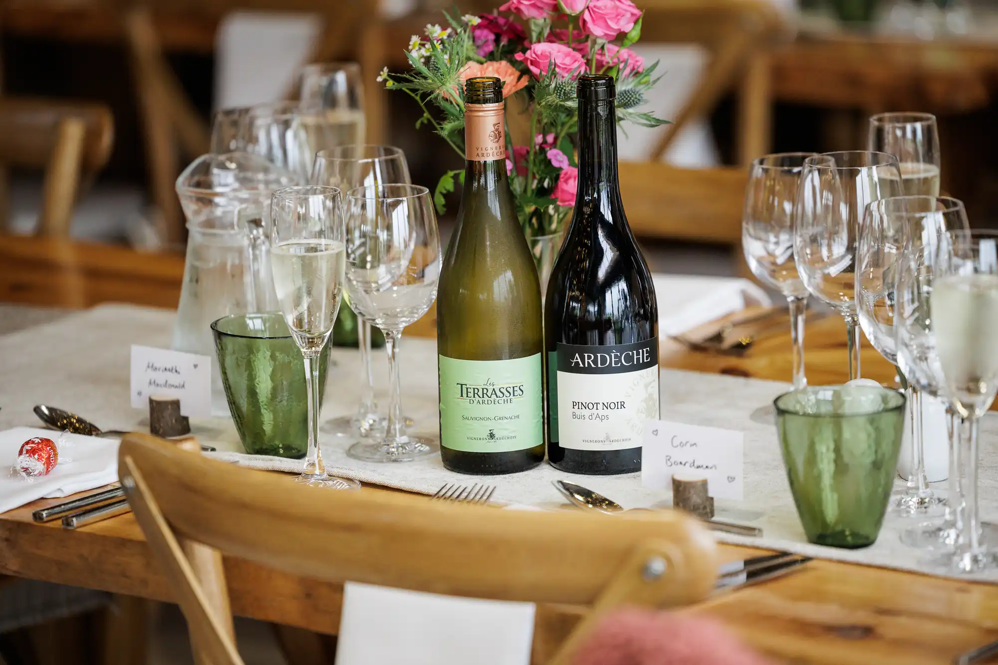 Two wine bottles (white and red) sit among wine glasses, water glasses, and flower arrangements on a wooden table laid for a meal. Pink flowers are in a vase. Wooden chairs and handwritten name cards are visible, creating a formal dining atmosphere.
