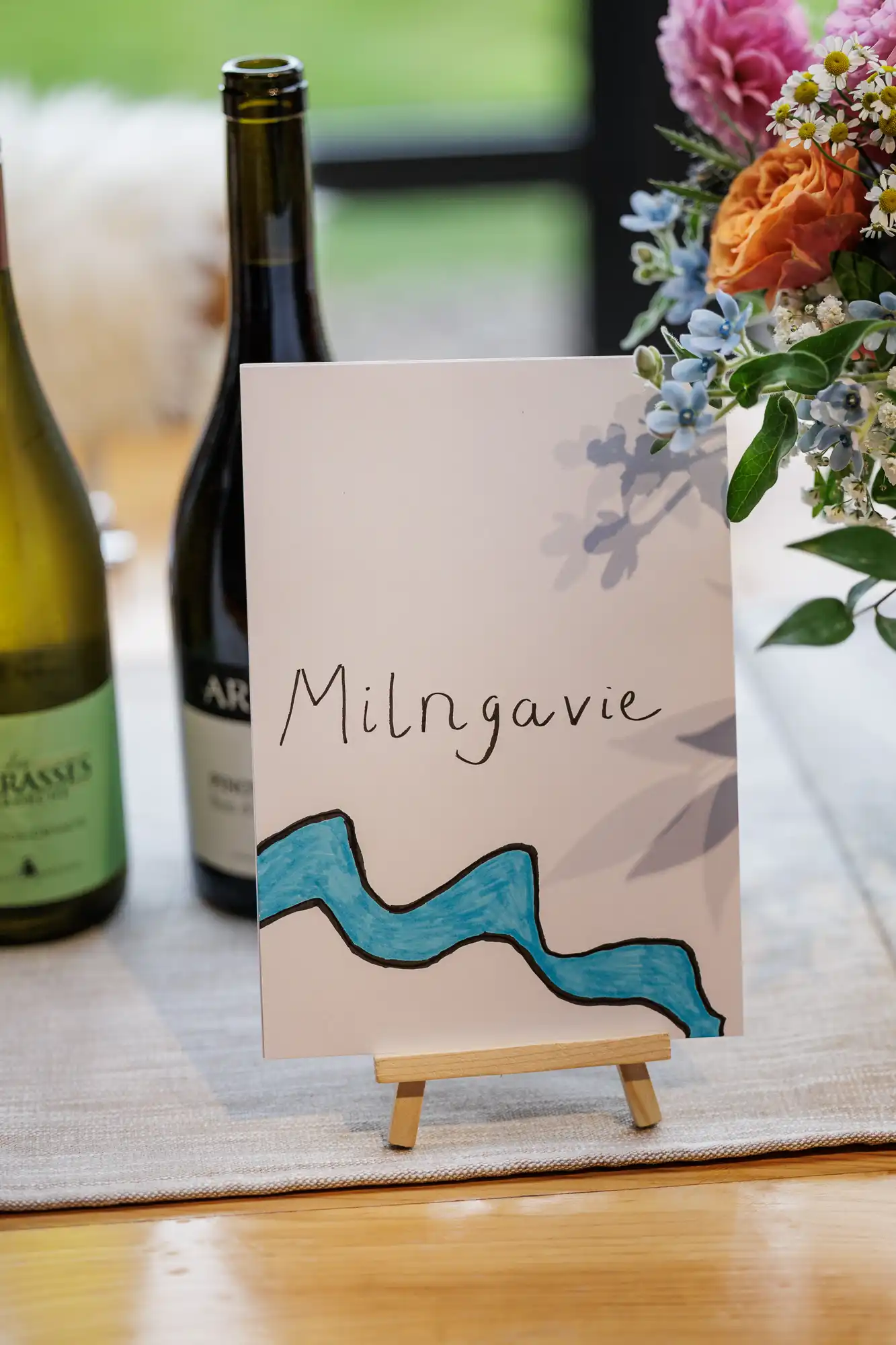 A white card with “Milngavie” handwritten in black and a blue wavy line is displayed on a small wooden easel. The card is on a table with wine bottles and a vase of pink and blue flowers in the background.