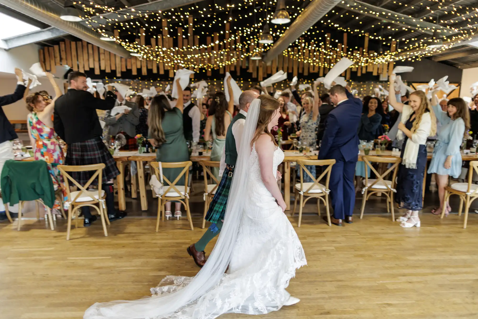 A bride and groom enter a wedding reception as guests stand, waving white serviettes. Fairy lights hang from the ceiling, creating a festive atmosphere. Wooden tables are decorated for the celebration. The bride wears a white dress and veil; the groom wears a kilt.