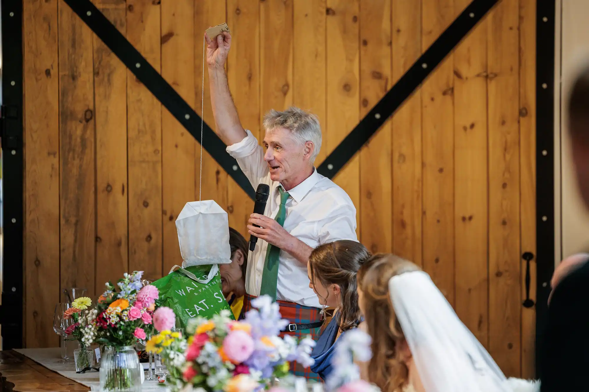 An older man in a white shirt and green tie stands at a table decorated with colourful flowers. He holds a paper bag in one hand and speaks into a microphone. Seated guests, including a bride in a veil, listen in front of a wooden barn-style door.