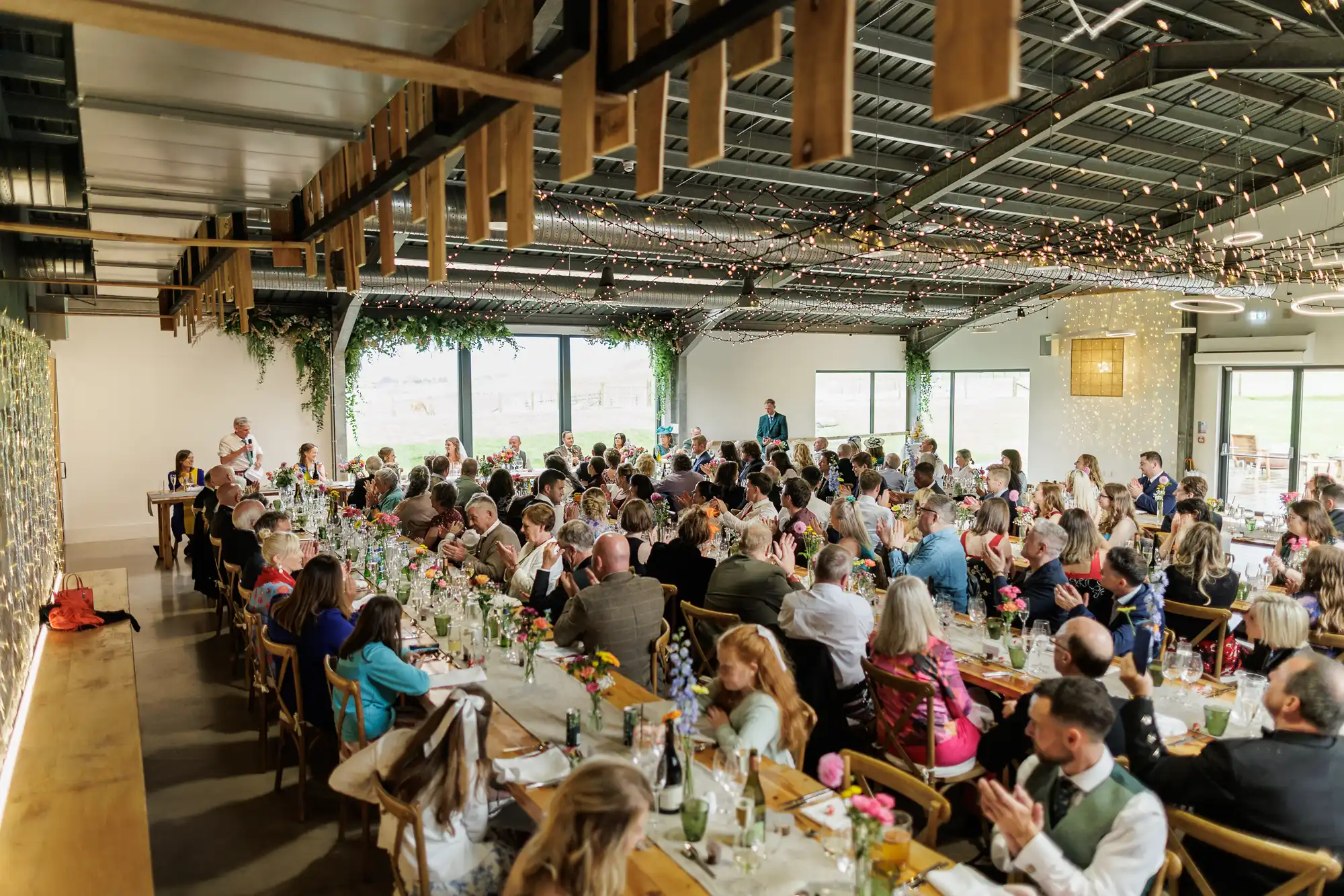 A large group of people sit at long, decorated tables inside a bright venue with large windows, greenery, and fairy lights on the ceiling. Guests face a speaker at the front, applauding, as they celebrate at what appears to be a wedding or formal event.