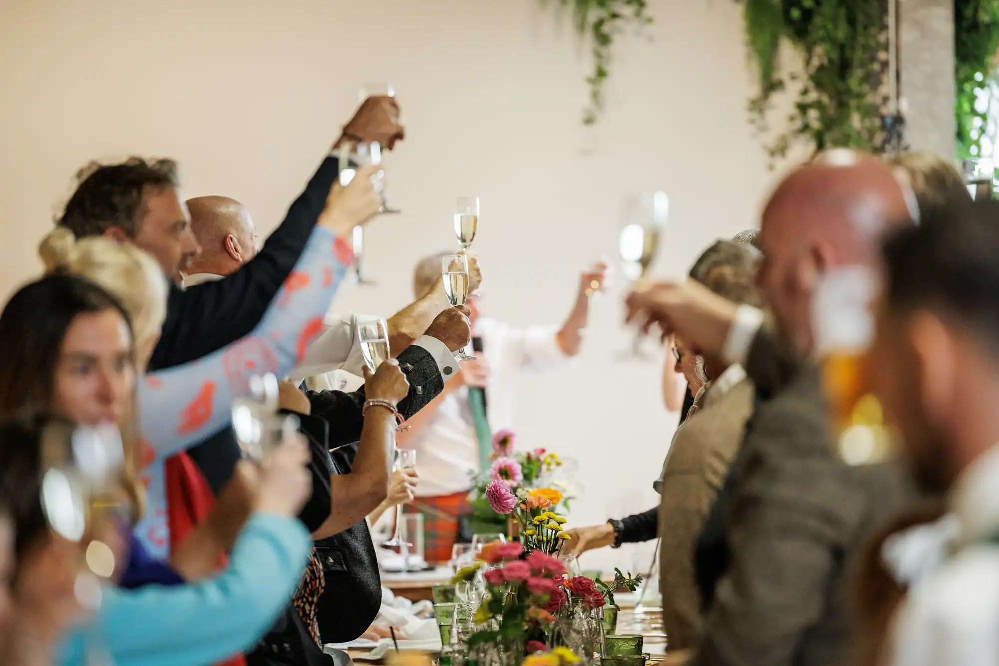 A group of people sit around a table raising glasses in a celebratory toast. The table is decorated with colourful flowers and greenery hangs on the wall in the background. The mood appears festive and joyful.