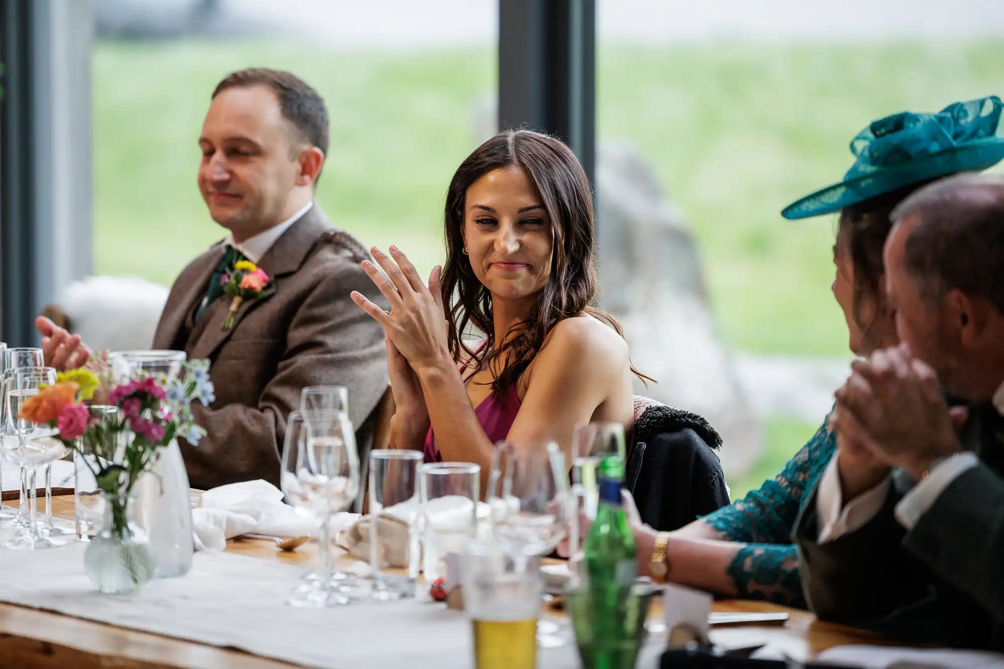 A woman in a pink dress smiles and claps at a table set for a formal meal, surrounded by others who are also clapping. There are wine glasses, flowers, and drinks on the rustic wooden table. Large windows with a green outdoor view are in the background.