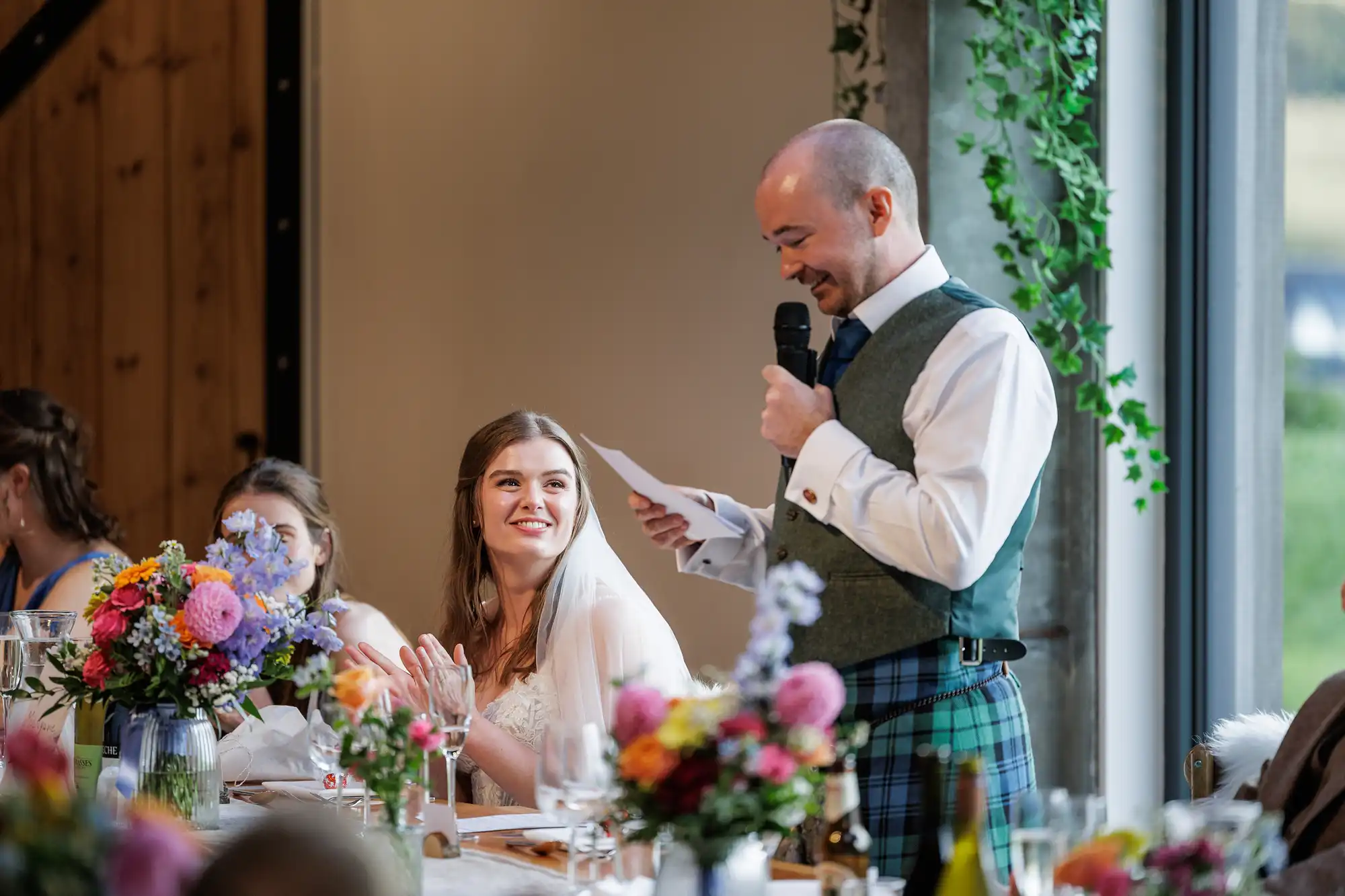 A groom in a kilt and waistcoat gives a speech with a microphone, smiling at his bride, who is seated beside him, wearing a white dress and veil, smiling and clapping. Colourful flower arrangements and guests surround them at a bright, decorated table.