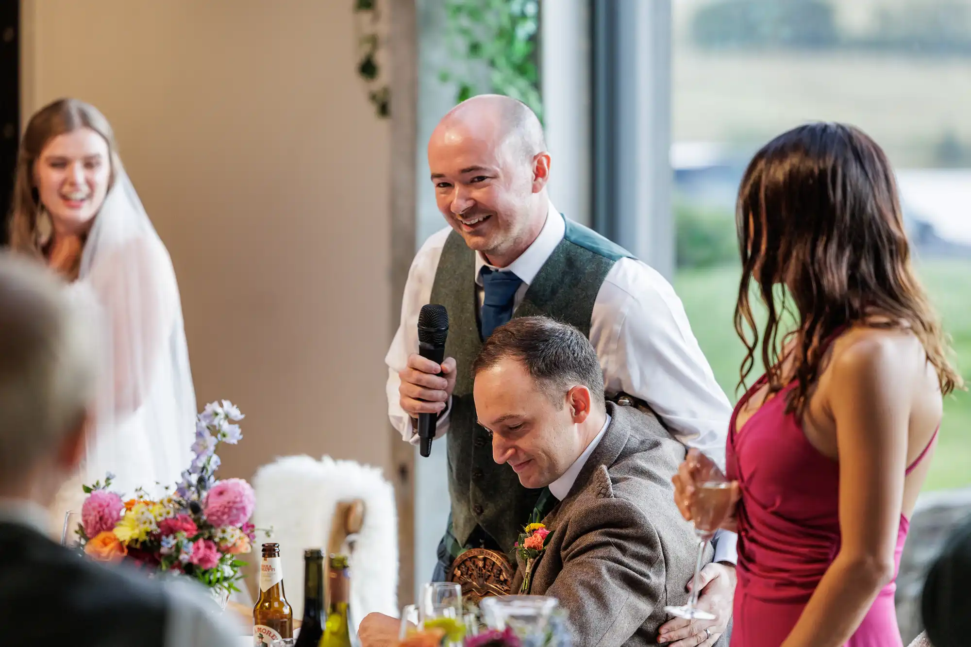A man in a green waistcoat stands holding a microphone, smiling, with his arm around a seated man in a suit. A woman in a pink dress stands next to them, and a bride in white looks on, all gathered round a table with colourful flowers and bottles.