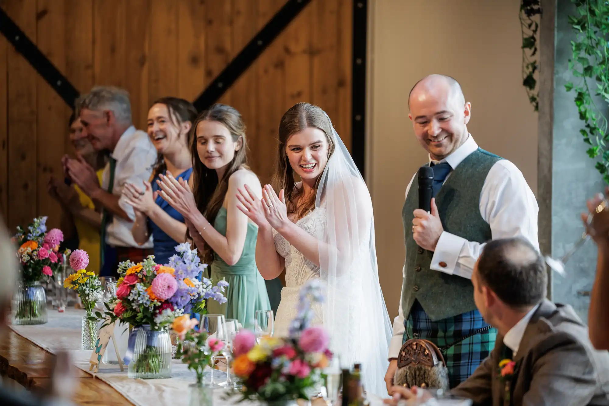 A bride in a white dress and veil smiles and claps at a decorated table, surrounded by applauding guests. A man in a waistcoat, kilt, and tie speaks into a microphone beside her. Colourful flowers and greenery decorate the bright, rustic setting.