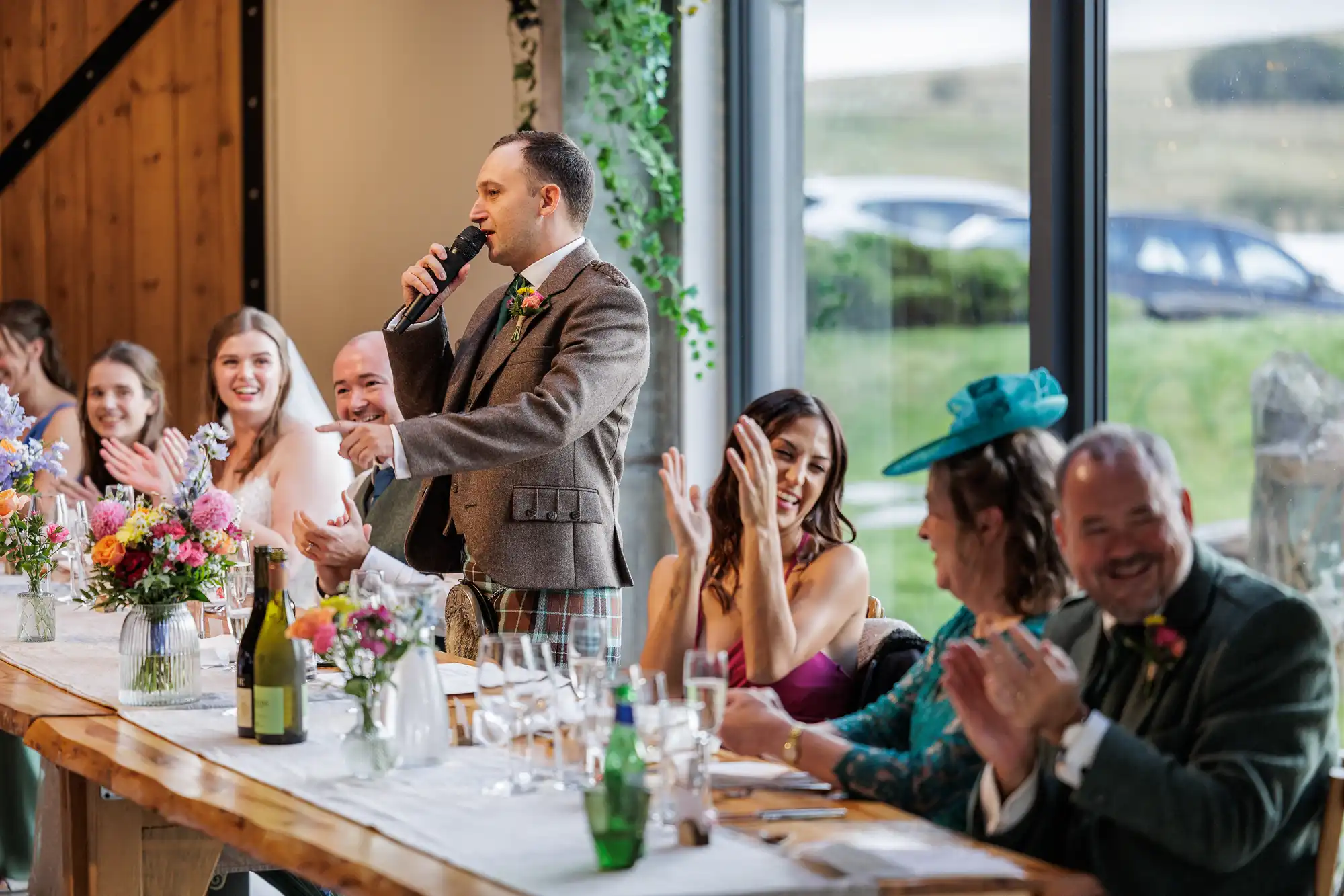 A man in a suit stands at a long table giving a speech with a microphone at a wedding reception. Guests seated on both sides, dressed formally, are smiling and applauding. The table is decorated with flowers and drinks; large windows show a scenic outdoor view.