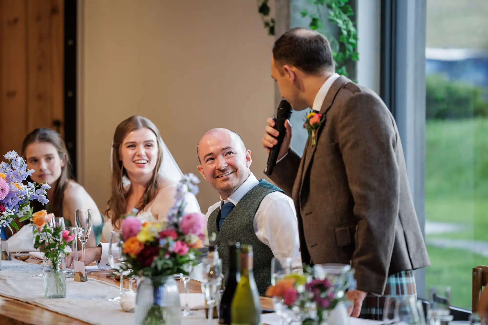 A man in a suit jacket stands and speaks into a microphone at a wedding reception table. The bride and groom sit smiling at him, with flower arrangements and wine bottles on the table. Two bridesmaids are also visible, and large windows show a green outdoor scene.