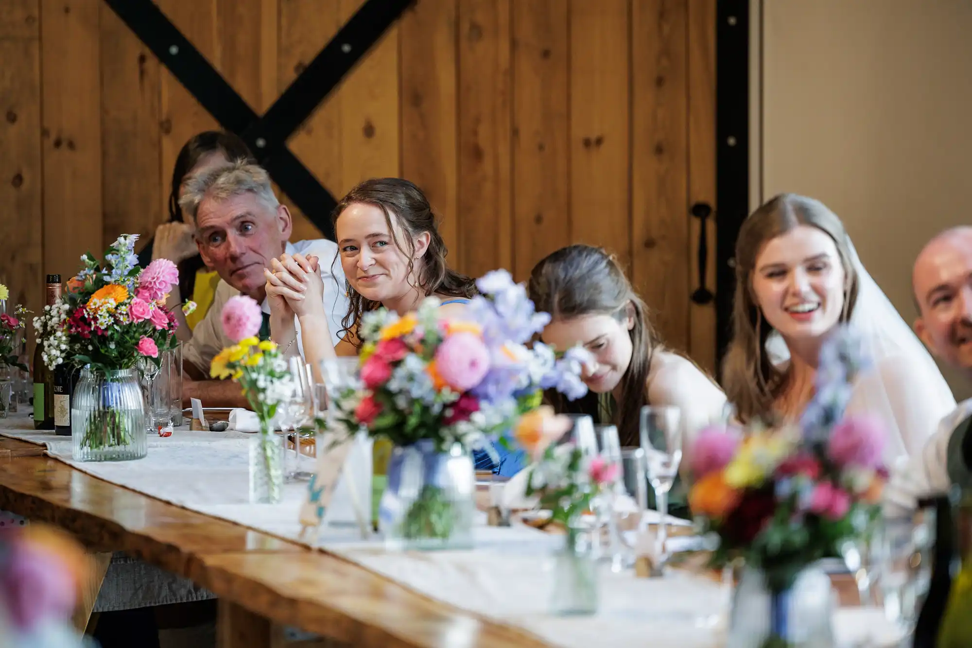 A group of people sit at a long table decorated with colourful flower arrangements. They are smiling and laughing, dressed in formal attire, with wooden walls in the background, suggesting a relaxed, joyous celebration or wedding reception.