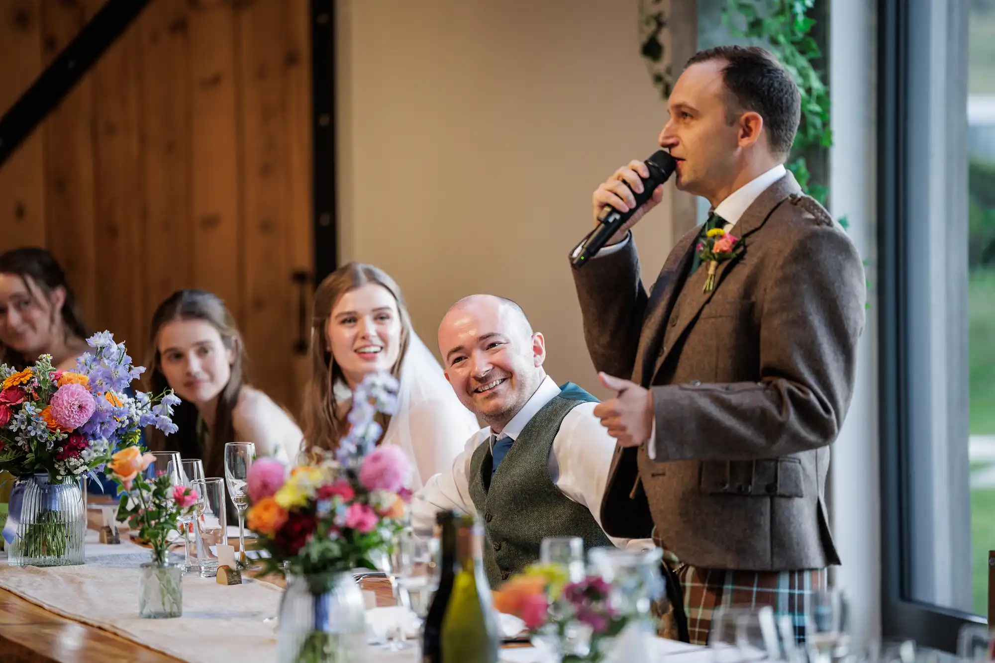 A man in a brown jacket speaks into a microphone at a wedding reception table, smiling guests listen and laugh. Colourful floral arrangements and glasses decorate the table, with natural light coming through large windows on the right.