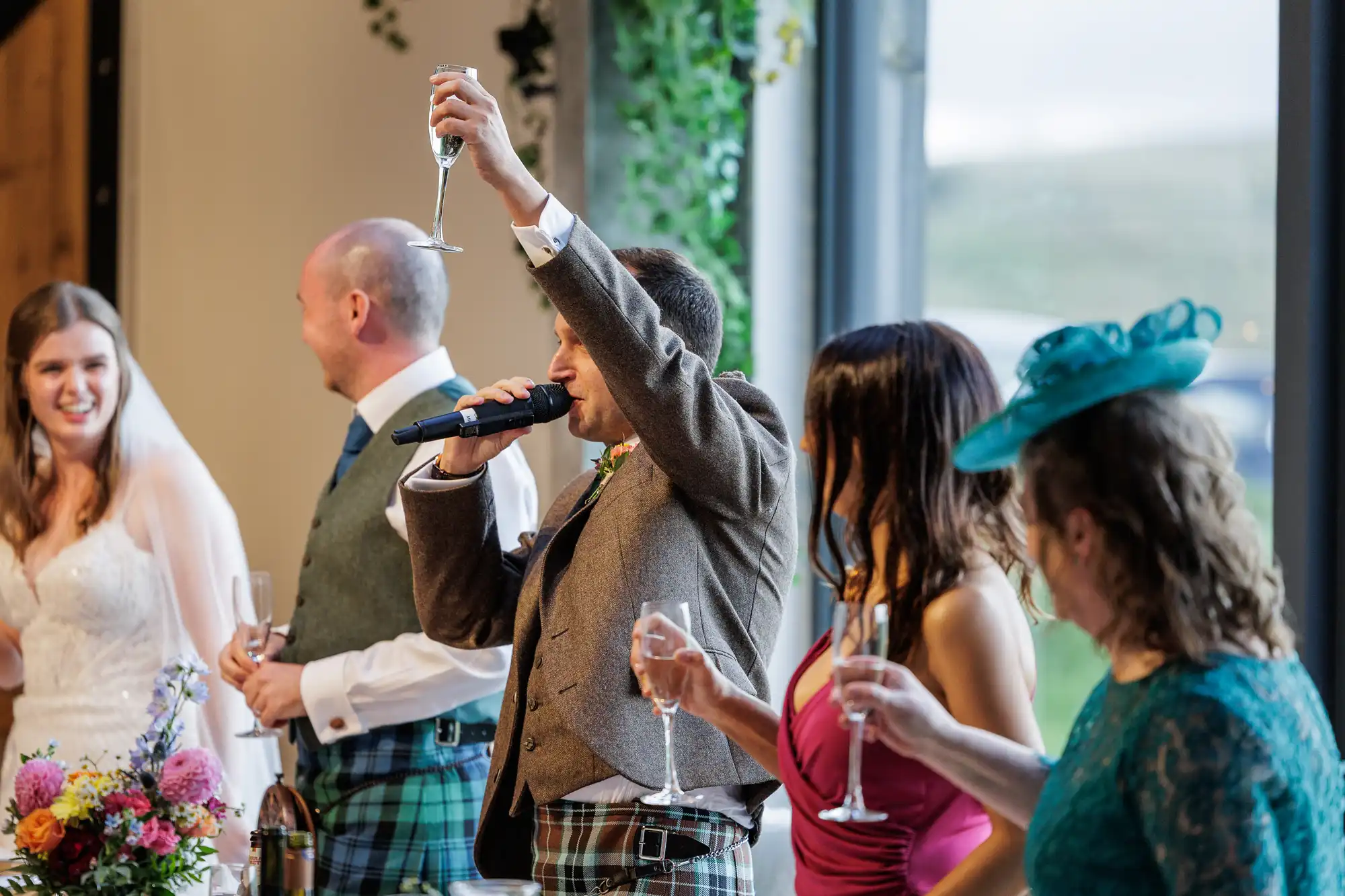A group of people at a wedding, all holding champagne glasses. A man in a tweed jacket and tartan trousers raises his glass and speaks into a microphone. A smiling bride stands nearby with three other guests, including a woman in a teal hat and dress.