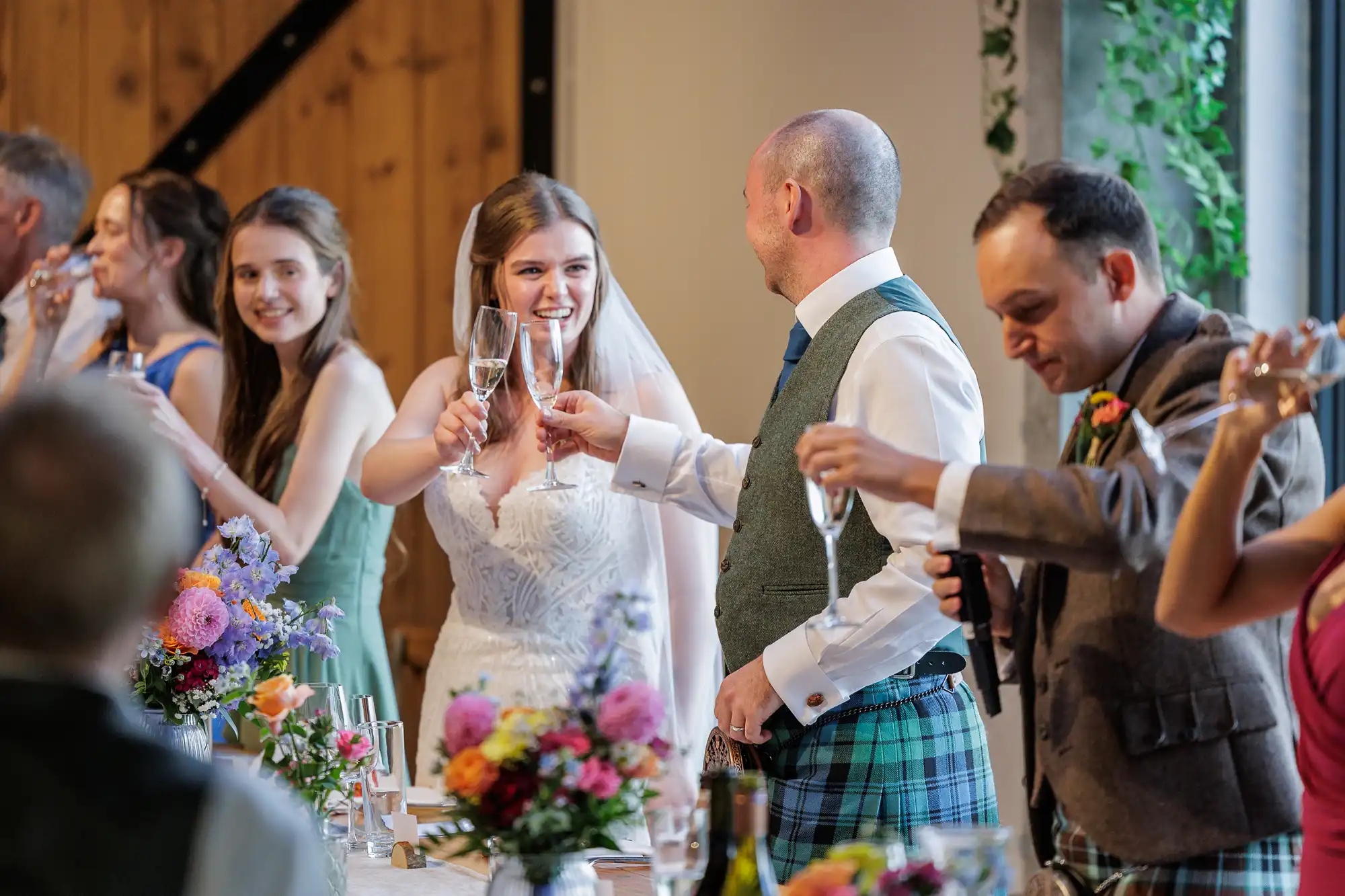A bride and groom in wedding attire raise champagne glasses for a toast, smiling at each other. Guests beside them also toast. The table is decorated with colourful flower arrangements, and there’s a warmly lit, festive atmosphere.
