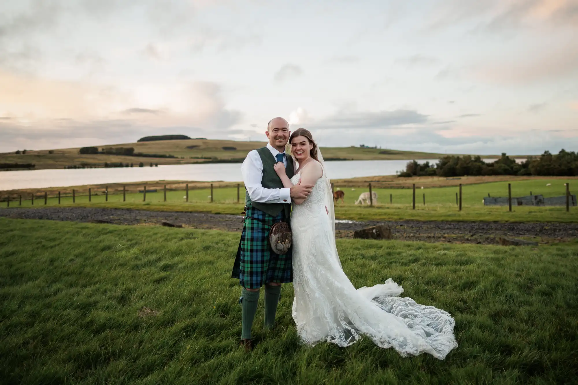 A bride in a white lace gown with a long train and veil stands beside a groom in a traditional Scottish kilt. They smile, embracing on green grass with a lake, rolling hills, and a cloudy sky in the background.