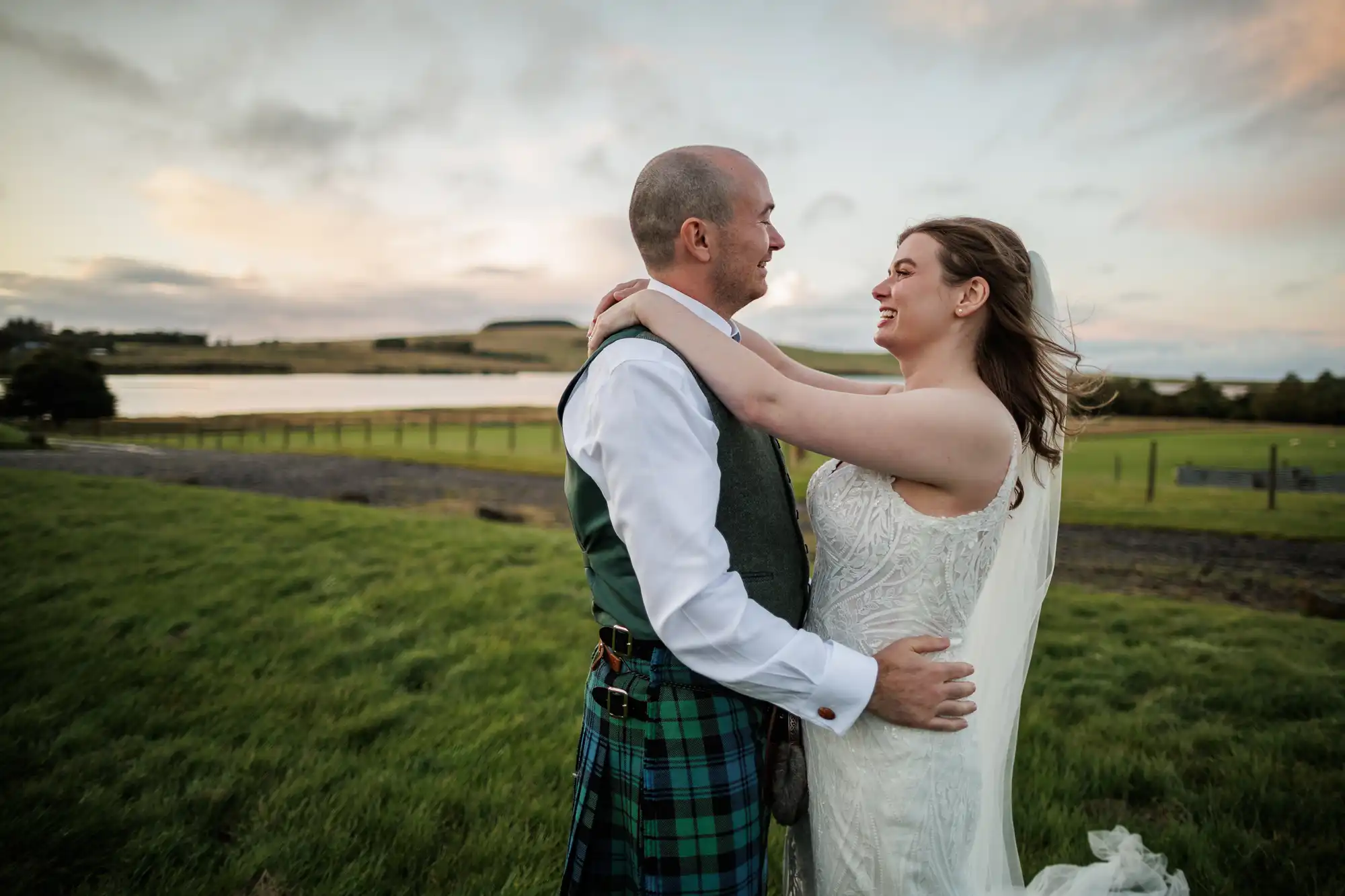 A bride in a white dress and a groom in a kilt embrace and smile at each other outdoors, with green fields, a fence, and a lake in the background under a partly cloudy sky at sunset.