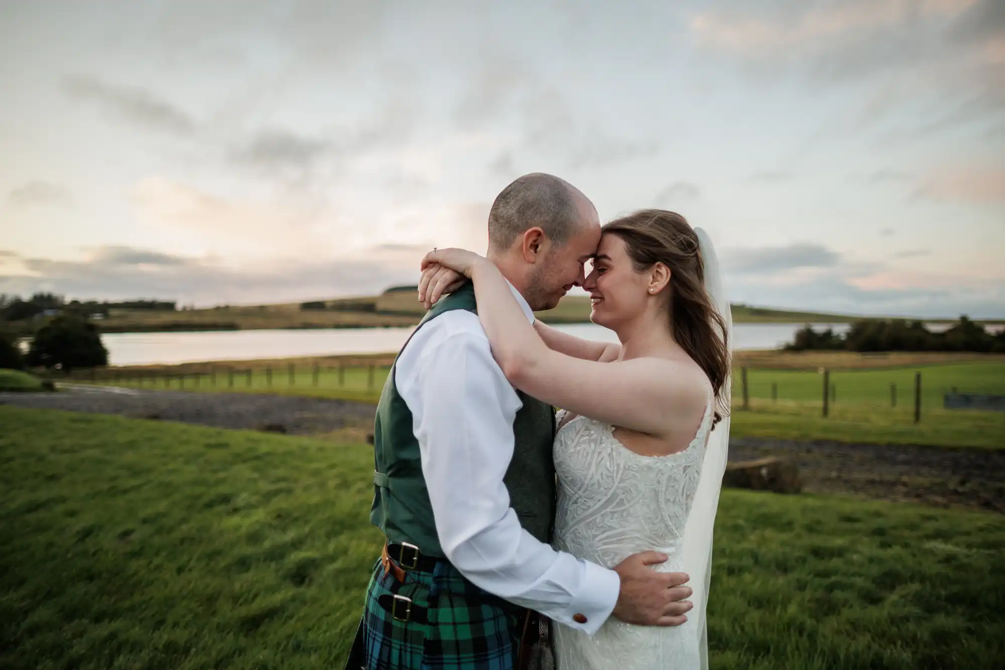 A bride in a white dress and veil embraces a groom in a white shirt, waistcoat, and tartan kilt. They stand closely together on green grass, forehead to forehead, with a scenic lake and cloudy sky in the background at sunset.